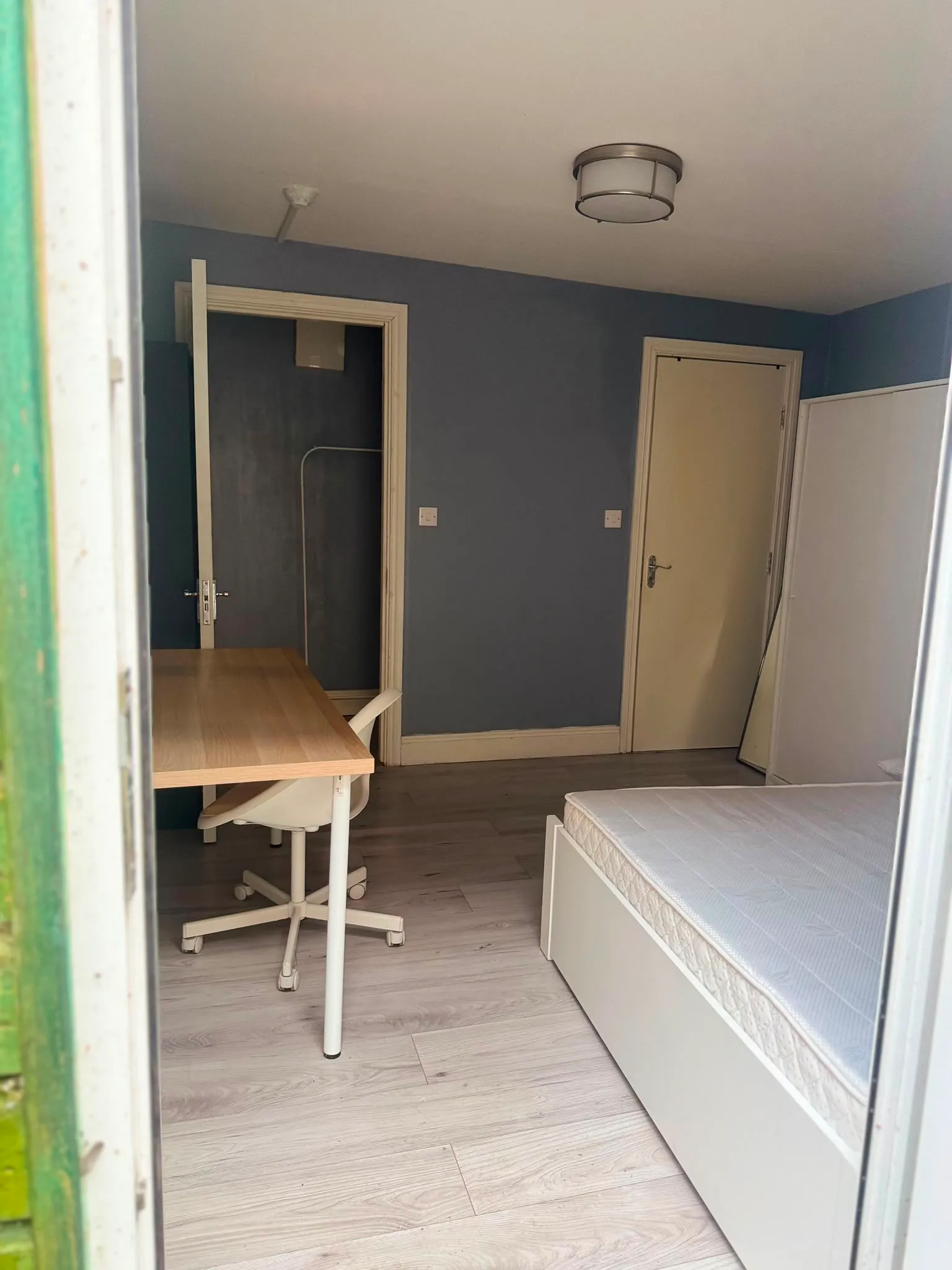 Bedroom with a single bed frame and mattress at right, a wooden desk and rolling chair at left, two doors and a ceiling light.