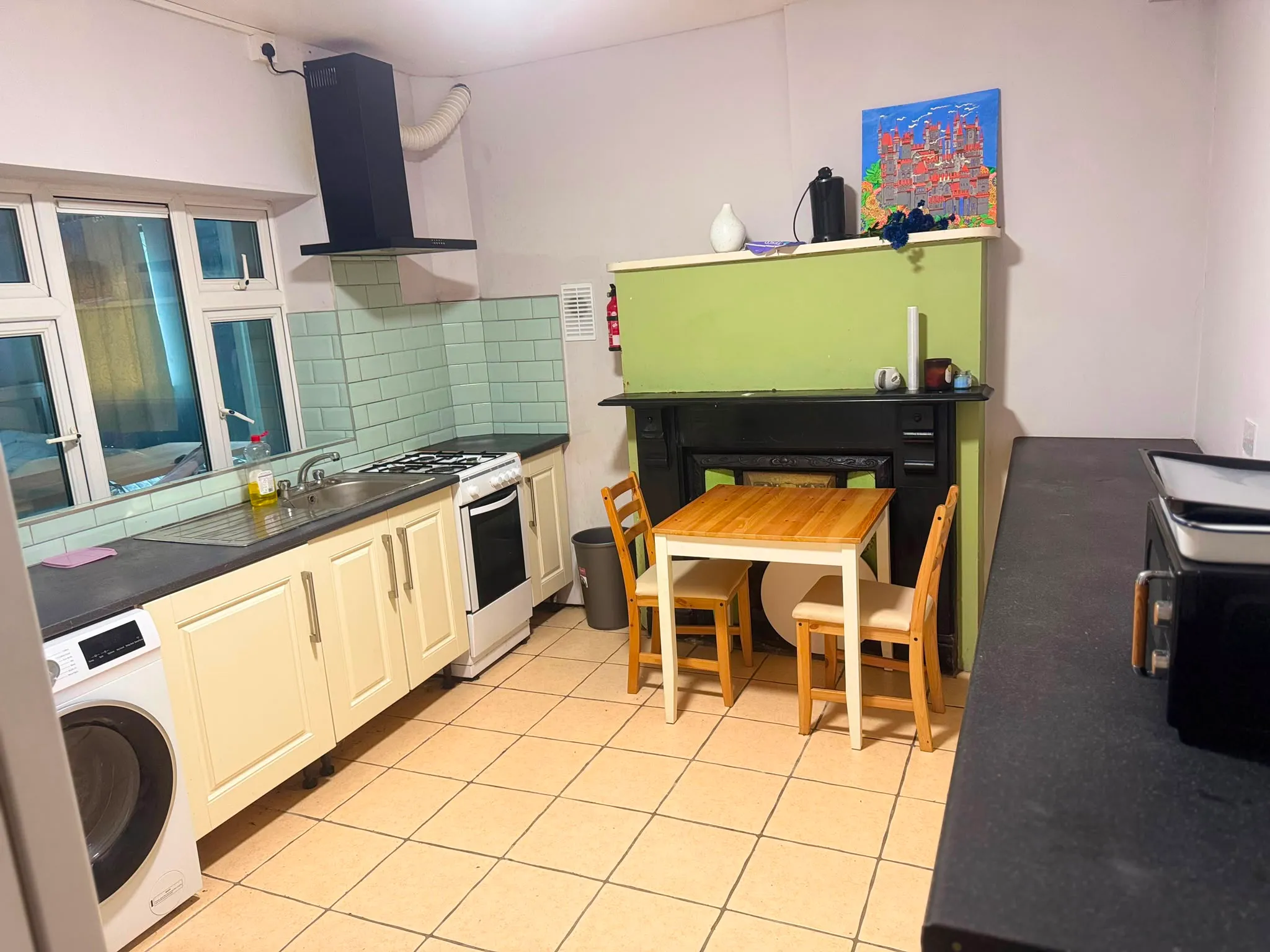 Kitchen with sink, gas stove and extractor hood on the left, a small wooden dining table with chairs in front of a fireplace, cabinets and windows above the sink.