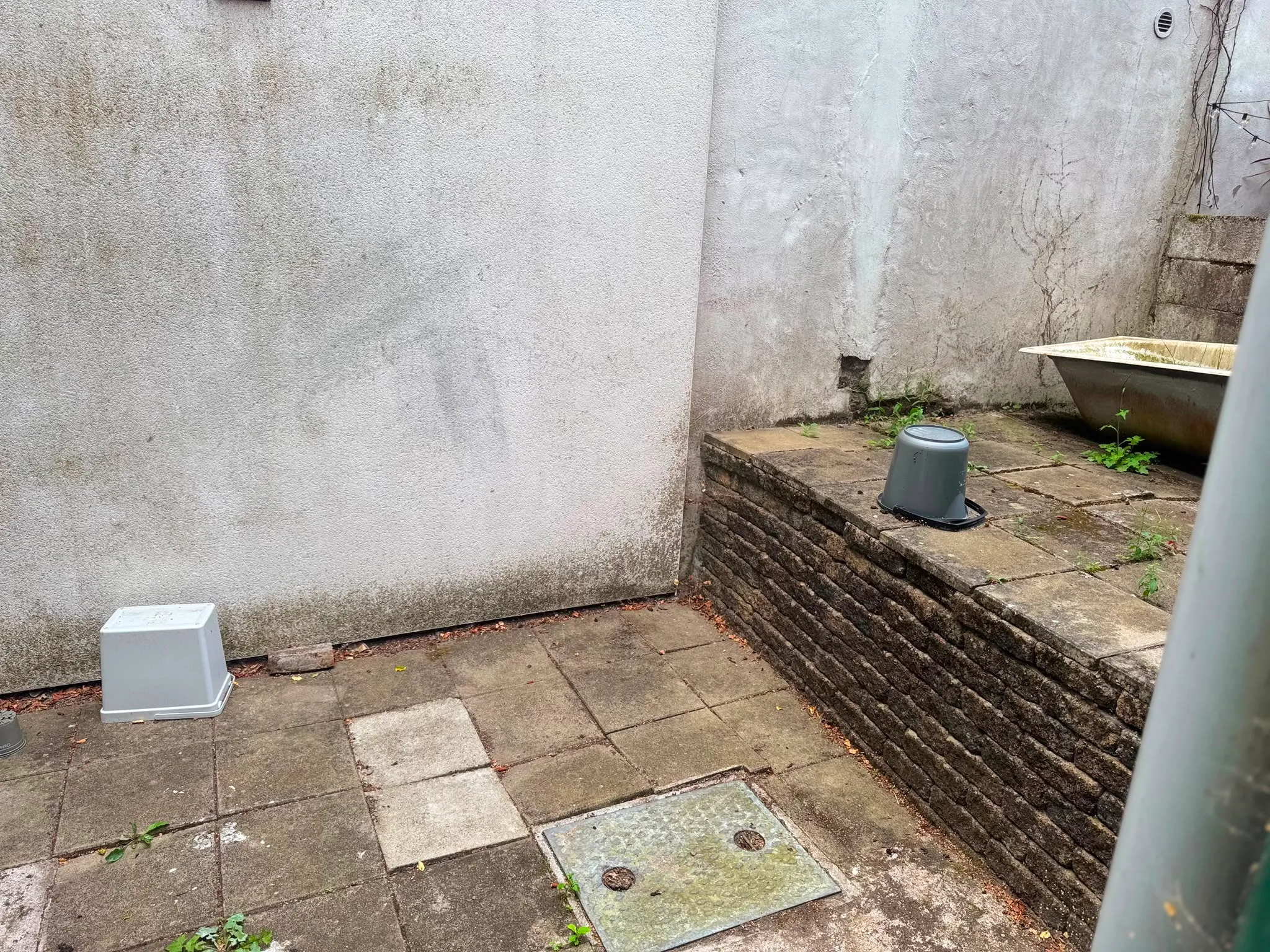 Small exterior paved courtyard with concrete walls, a low brick retaining wall, an upturned plastic bucket and an old bathtub at the edge
