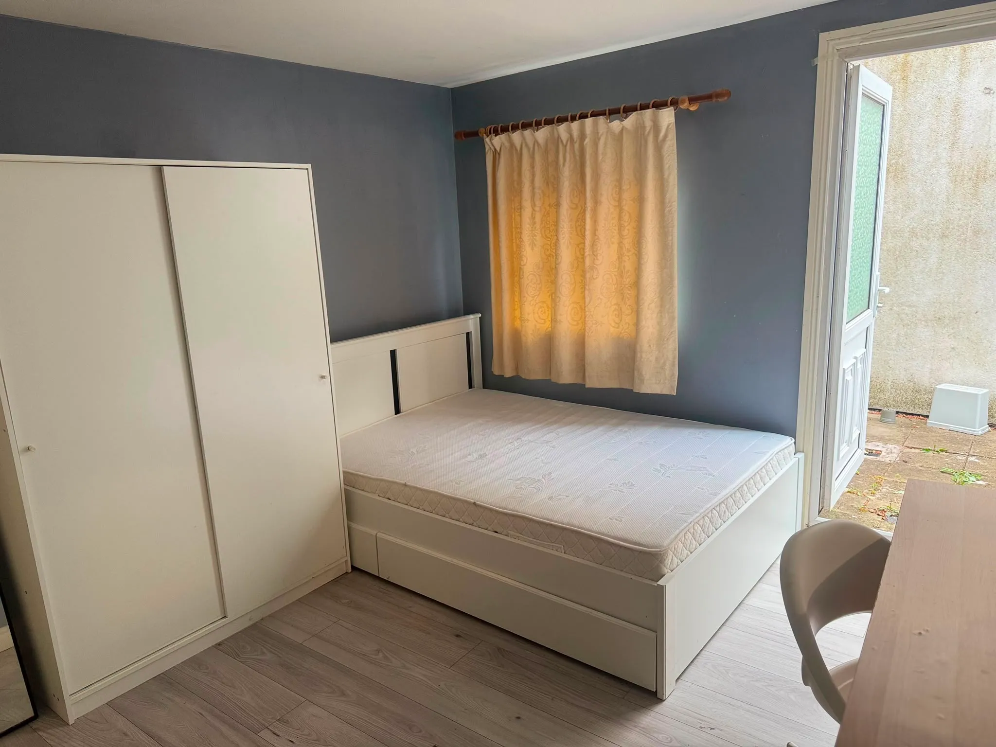 Bedroom with a double bed frame and mattress, white wardrobe on the left, a curtained window, and a door opening to an outdoor area; a desk and chair visible at right.