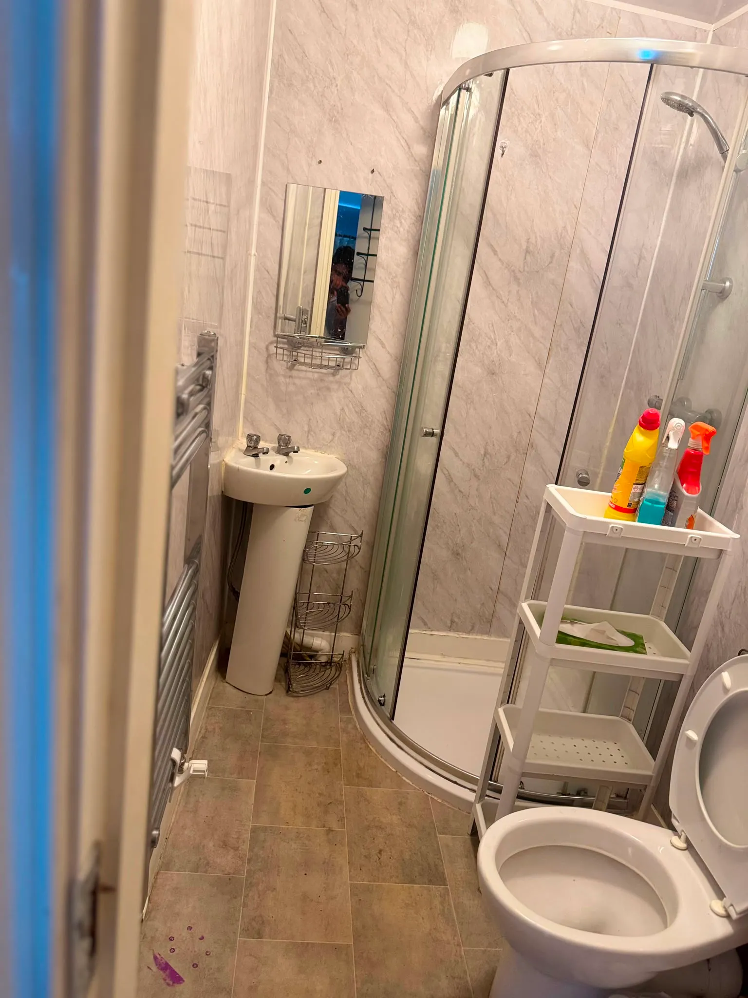 Small bathroom with a corner glass shower, pedestal sink with wall mirror, toilet with open seat, and a three-tier plastic storage cart holding cleaning bottles.