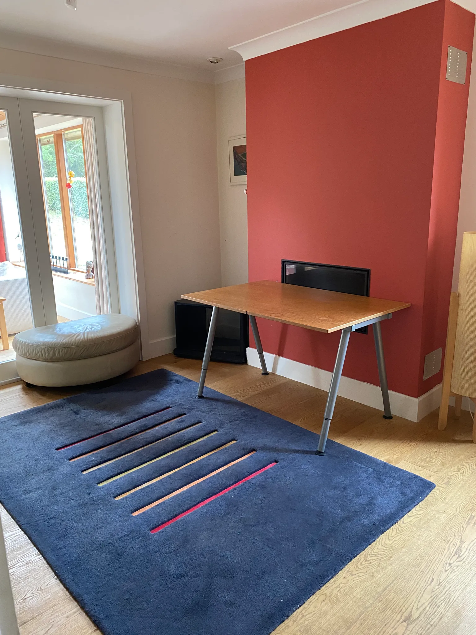 Living room with a small wooden table against a red accent wall, a blue rug on a wood floor, a round ottoman and glass doors leading outside.