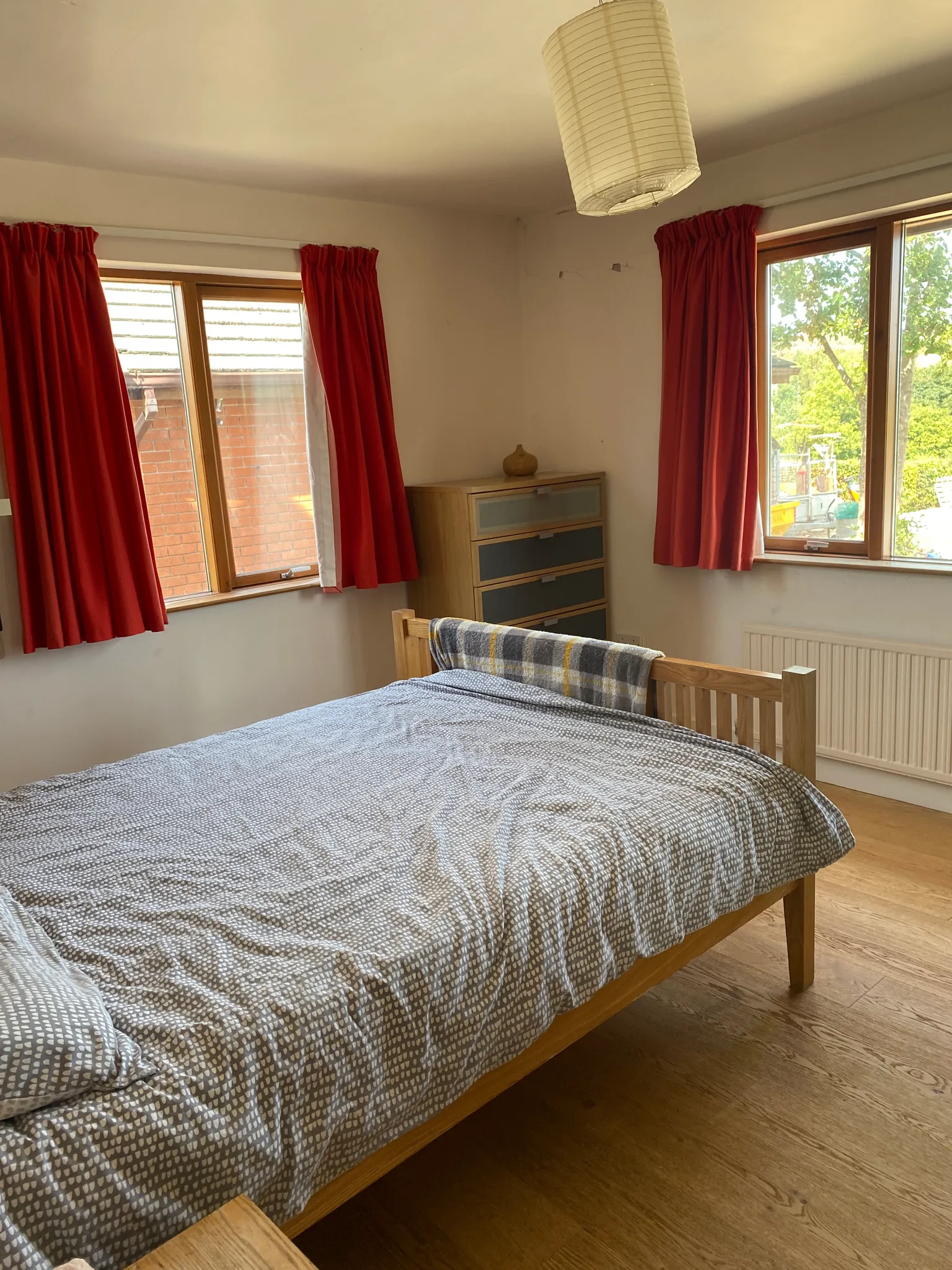 Bedroom with a wooden double bed, patterned bedding, two windows with red curtains, a chest of drawers and a hanging ceiling lamp.