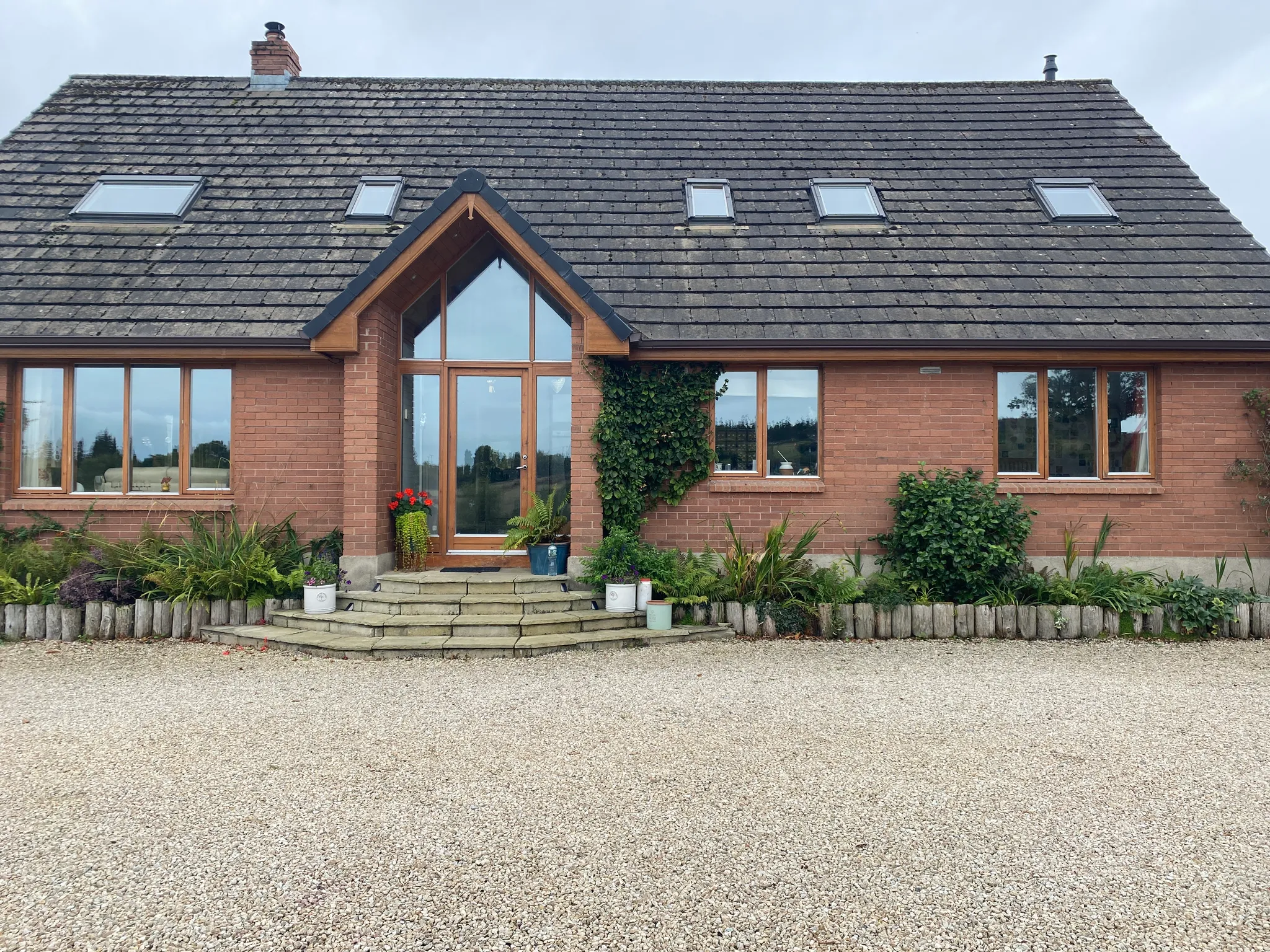 Exterior front view of a brick house with a steep tiled roof, four skylights, a central glass door with triangular window above, front steps and planted borders