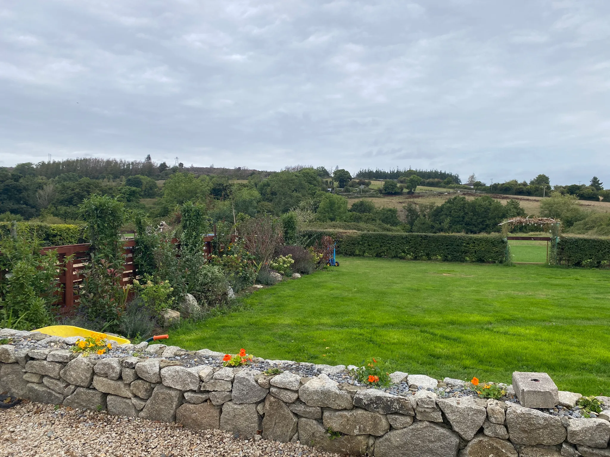 Back garden with a green lawn, low stone wall and hedge with a gate opening to fields and wooded hills under a cloudy sky.