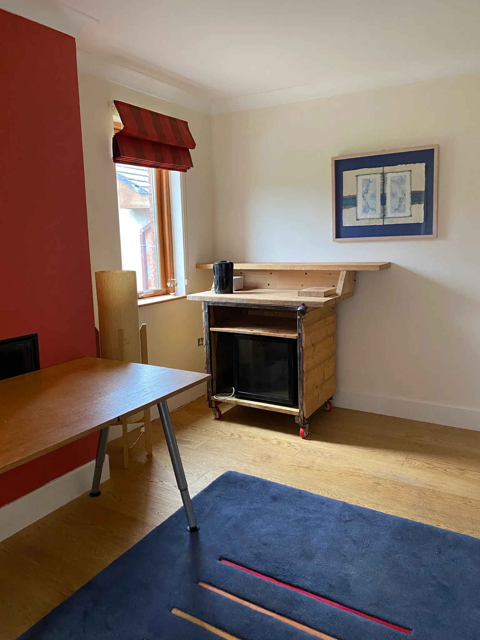 Small workspace with a wooden desk, rolling wooden cabinet with a mini fridge, window with a red roman blind, framed artwork on the wall and a blue rug.