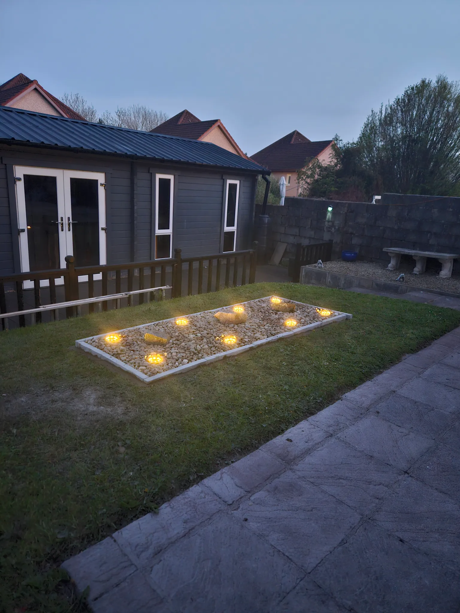 Backyard exterior with a small dark-gray shed with white doors and windows, a gravel planting bed with round lights, low stone bench and paved path.