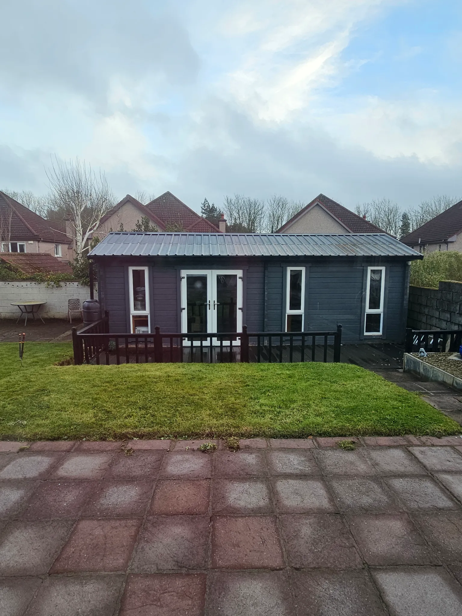 Backyard exterior showing a detached grey wooden outbuilding with double glass doors and side windows, small raised deck with railing, a lawn in front, paved area in foreground and neighboring houses visible behind.