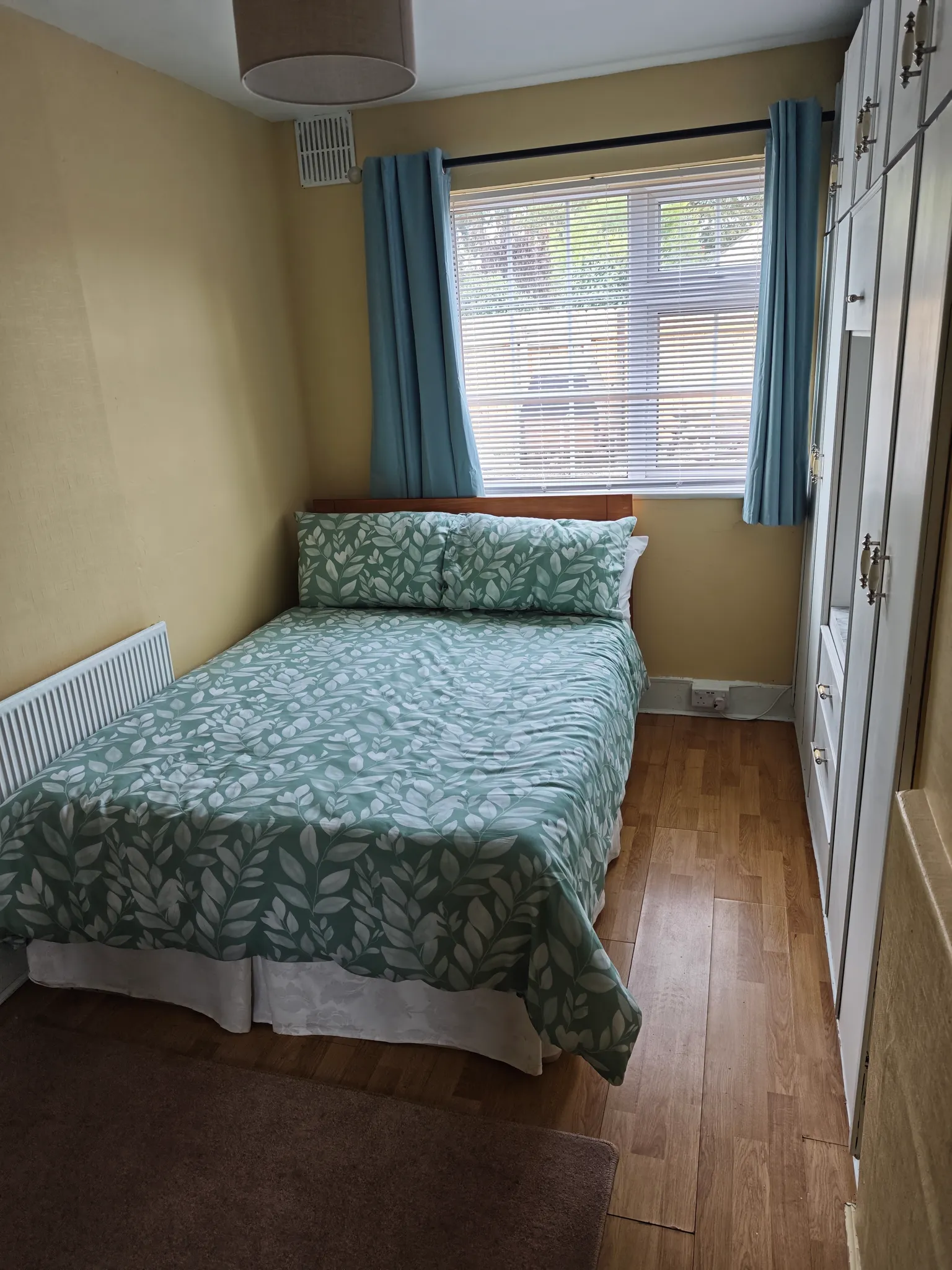 Bedroom with a double bed with patterned duvet, window with blinds and blue curtains, built-in wardrobe along one wall and radiator.