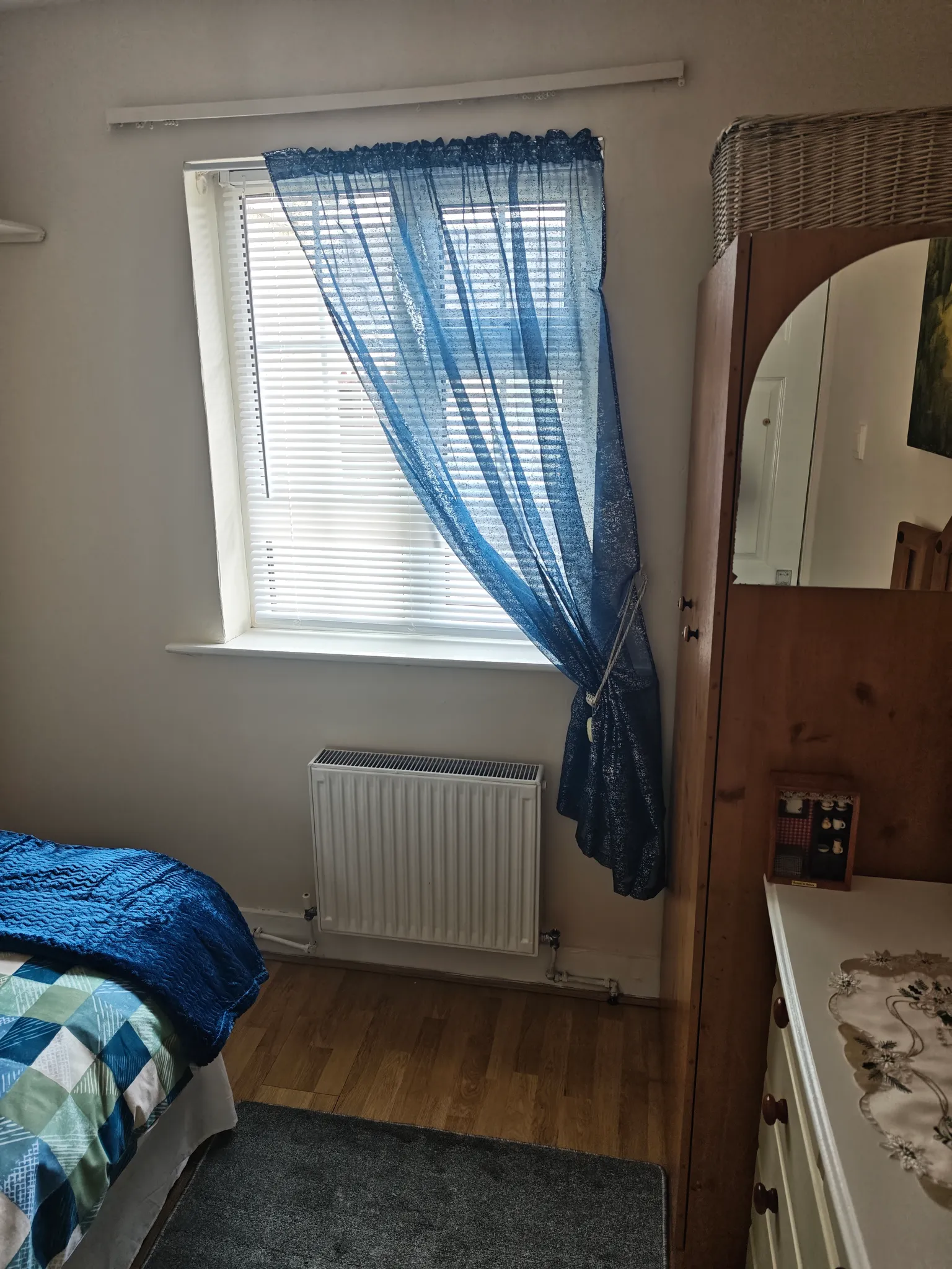 Bedroom corner with a window fitted with white blinds and a sheer blue curtain tied to one side, a radiator below the window, part of a bed with blue bedding at left, and a wooden wardrobe with mirror at right.