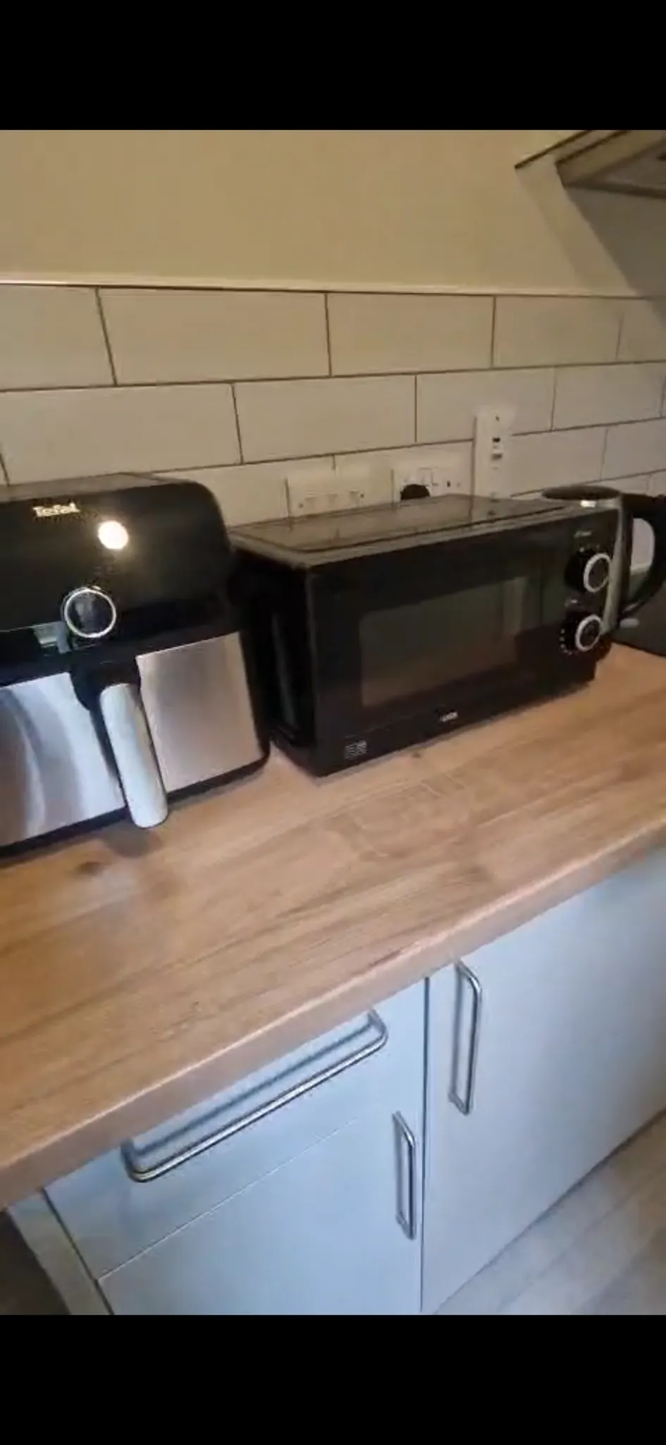 Kitchen countertop with a microwave, an air fryer and an electric kettle on a wood-effect counter with white tiled backsplash and cabinets below.