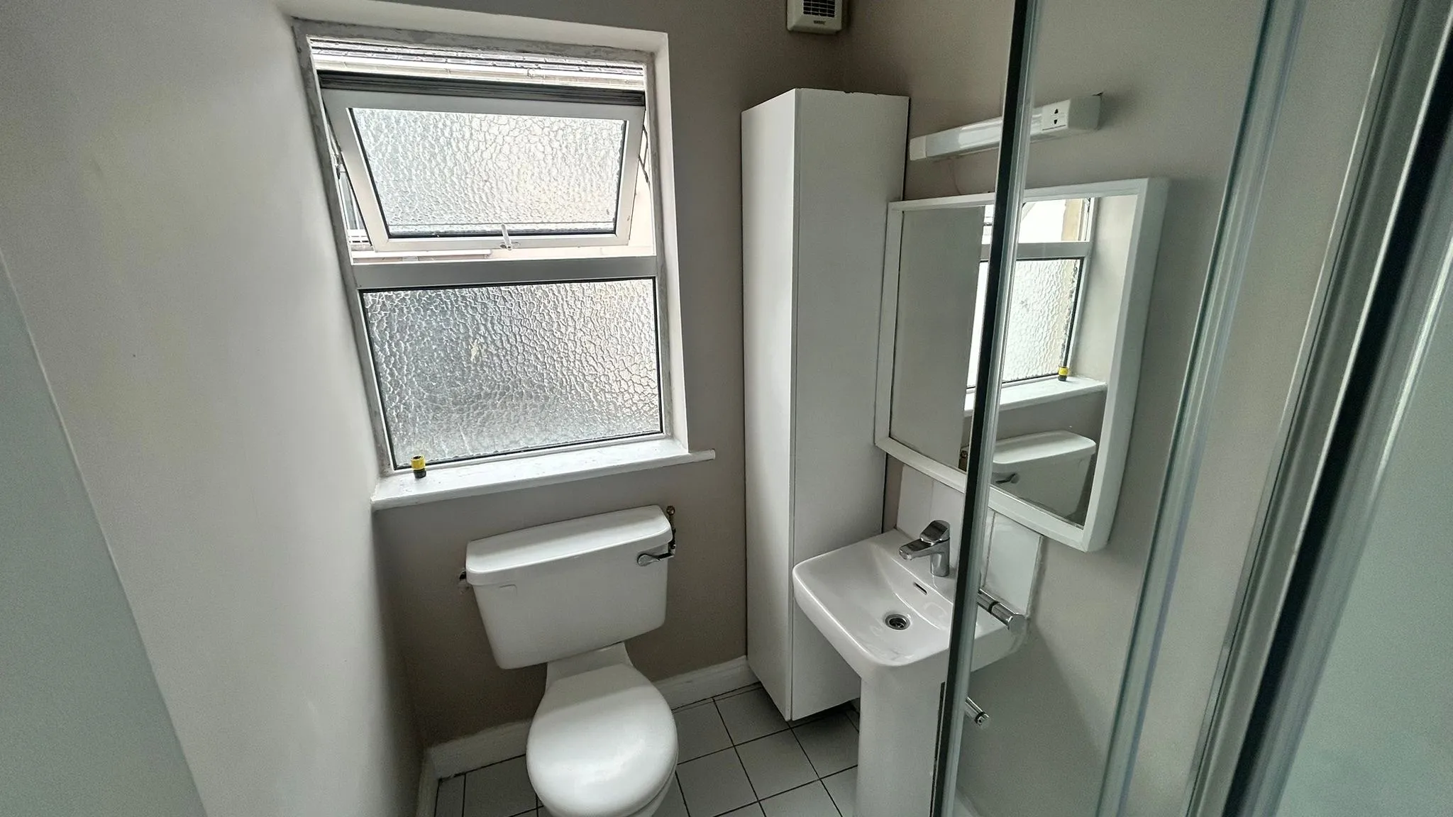 Small bathroom with a toilet, pedestal sink, mirrored cabinet, tall storage cupboard, frosted window, and a glass shower door.
