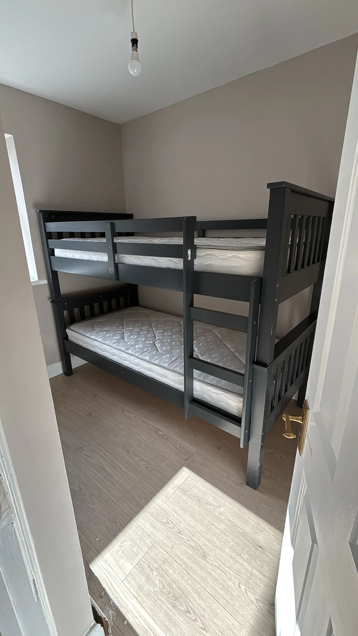 Small bedroom with a dark wooden bunk bed, two mattresses, a narrow window, bare ceiling bulb and light wood floor.