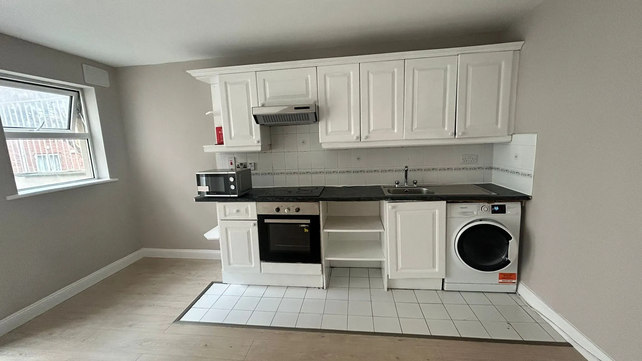 Kitchen area with white wall cabinets, electric oven and hob, microwave on the counter, sink, and a washing machine, with a window on the left.