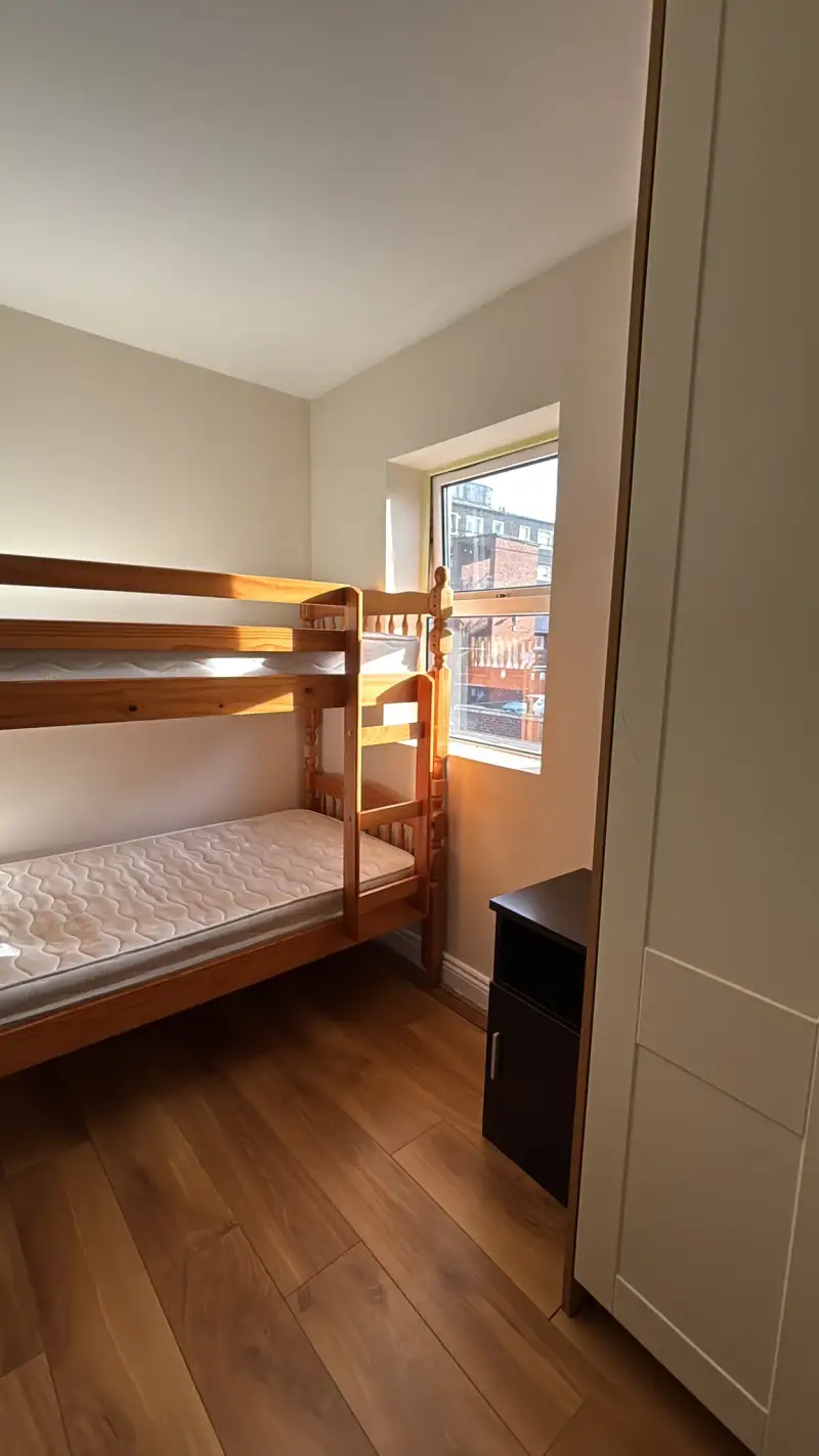 Bedroom with a wooden bunk bed, mattresses, a small window letting in daylight, wood floor and a partial view of a wardrobe and bedside cabinet.