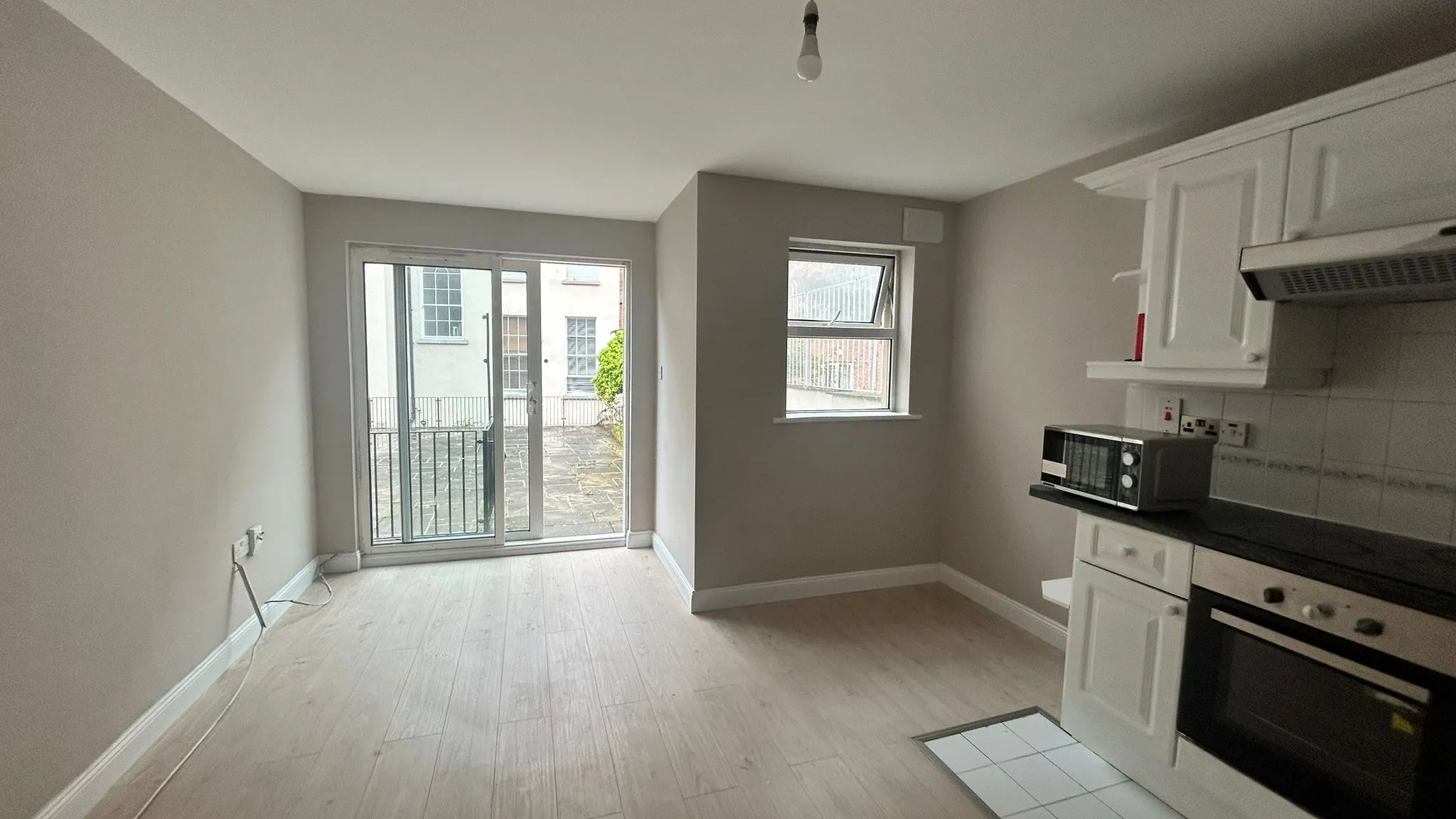 Open-plan room with a small kitchenette on the right, sliding glass doors to an exterior area, a small window, and light-colored wood-look flooring.