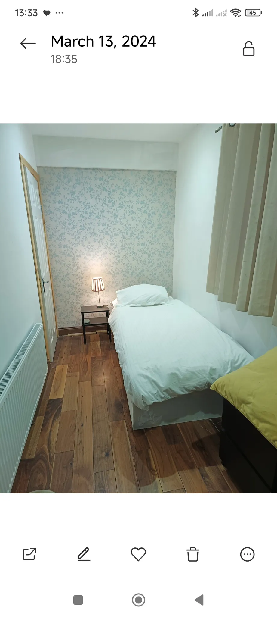 Bedroom with a single bed, bedside table with a lamp, patterned wallpaper on the back wall, curtains, wooden floor and a radiator.