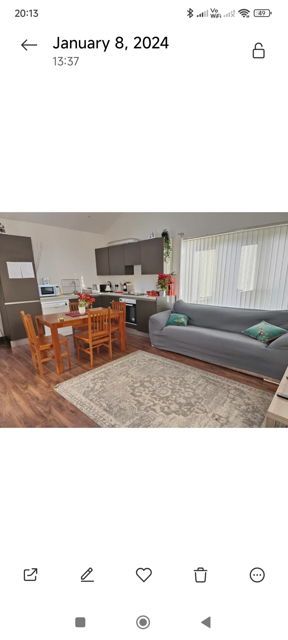 Open-plan living room and kitchen with a gray sofa, wooden dining table with four chairs, kitchen cabinets and appliances, large window with vertical blinds, and area rug on wood-look floor.