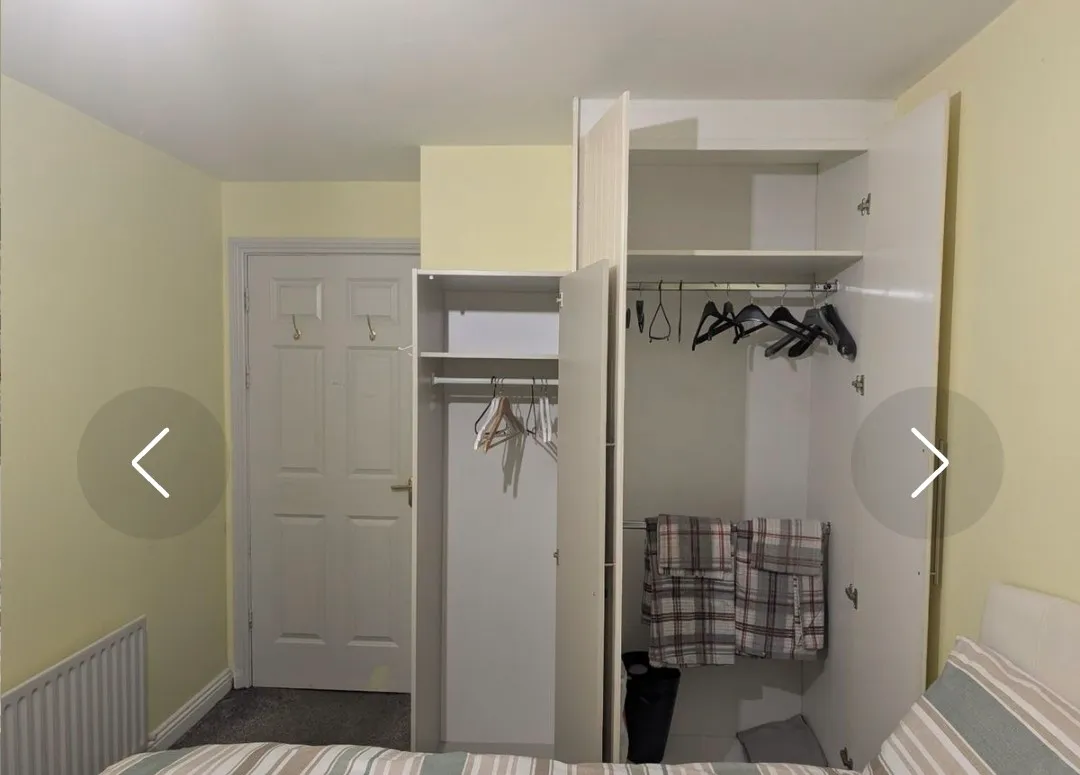 Bedroom corner showing an open built-in wardrobe with hangers and folded towels, a partially visible bed with striped bedding, a closed door and a radiator.