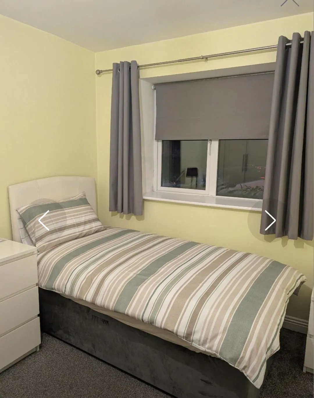 Bedroom with a single bed with striped duvet, pillow, bedside drawers, and a window with a gray roller blind and matching curtains.
