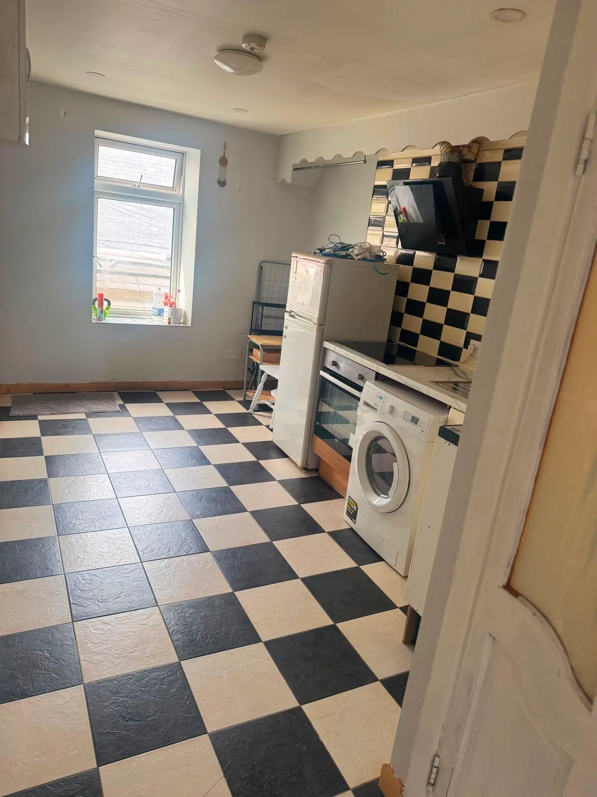 Kitchen with black-and-white checkered floor, washing machine, fridge, oven and a single window.
