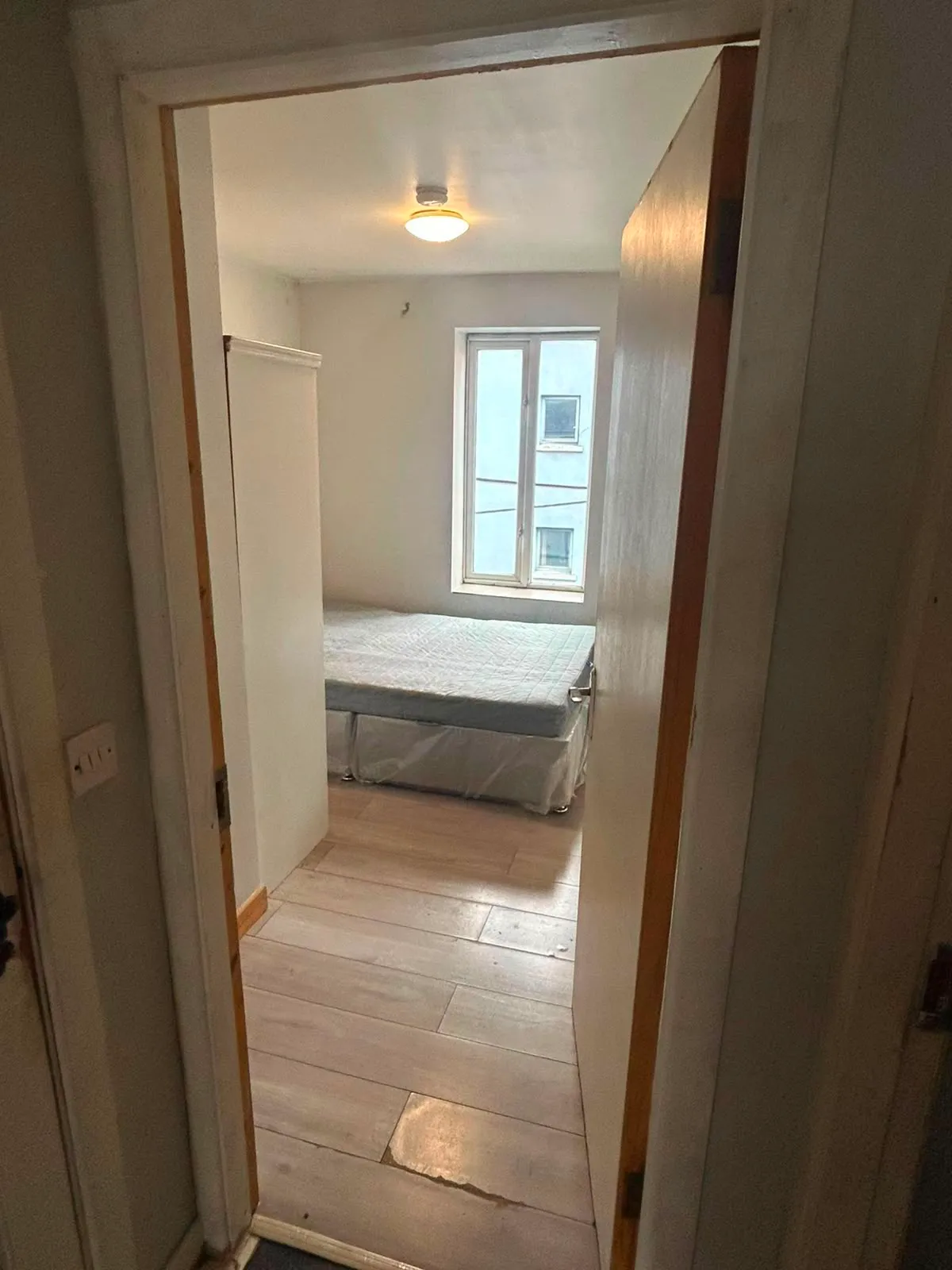 View through a doorway into a small bedroom with a mattress on a bed base, a narrow window on the far wall, a ceiling light, and a wardrobe partially visible on the left.