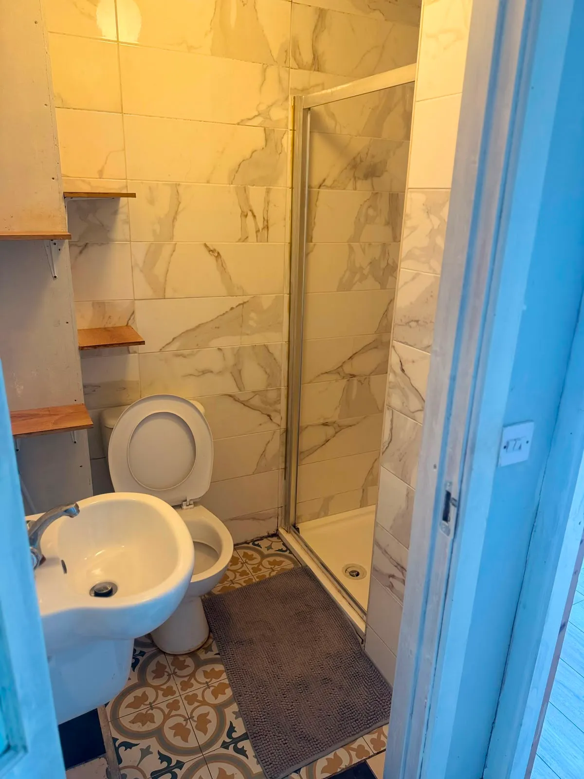 Small bathroom with a toilet (seat up), pedestal sink, walk-in shower with sliding door, wall-mounted shelves, and patterned floor tiles.