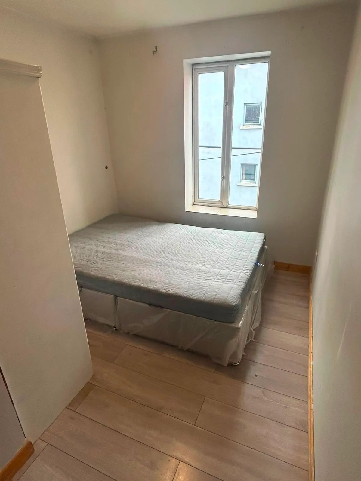 Small bedroom with a mattress on a bed base placed against the left wall, a tall narrow window on the far wall, light wood floor and plain white walls.