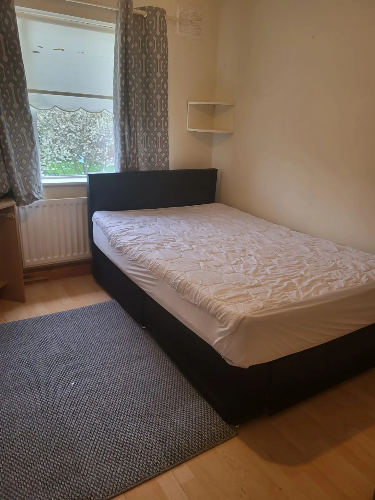 Bedroom with a double bed against a wall, window with patterned curtains, small wall shelf, radiator under the window and a rug on a wood floor.