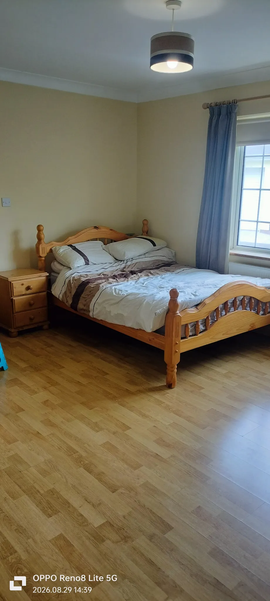 Bedroom with a wooden double bed, bedside table, window with curtain, ceiling light, and wood-look floor.