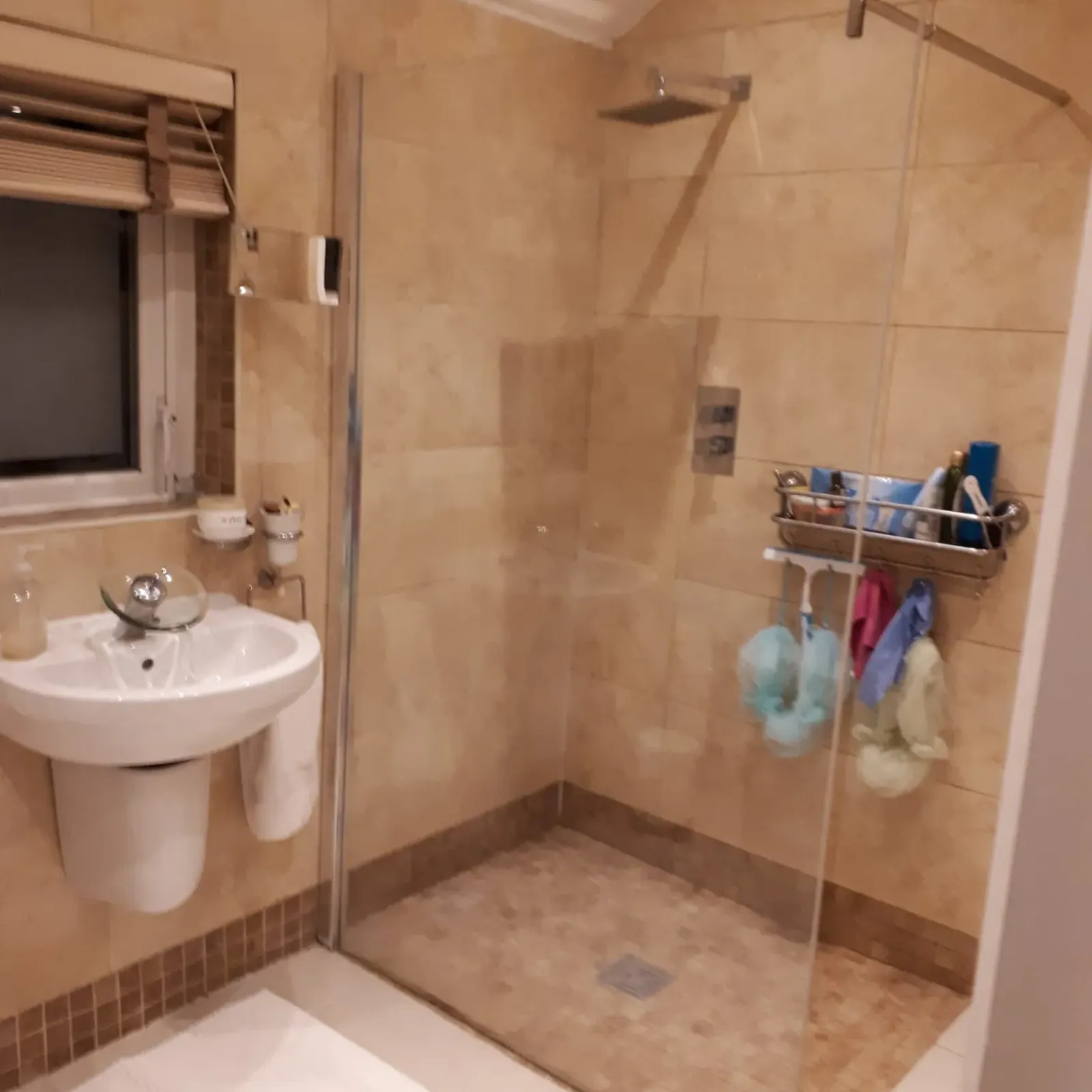 Bathroom with a walk-in tiled shower with glass partition, wall-mounted rain showerhead, pedestal sink and a small window.