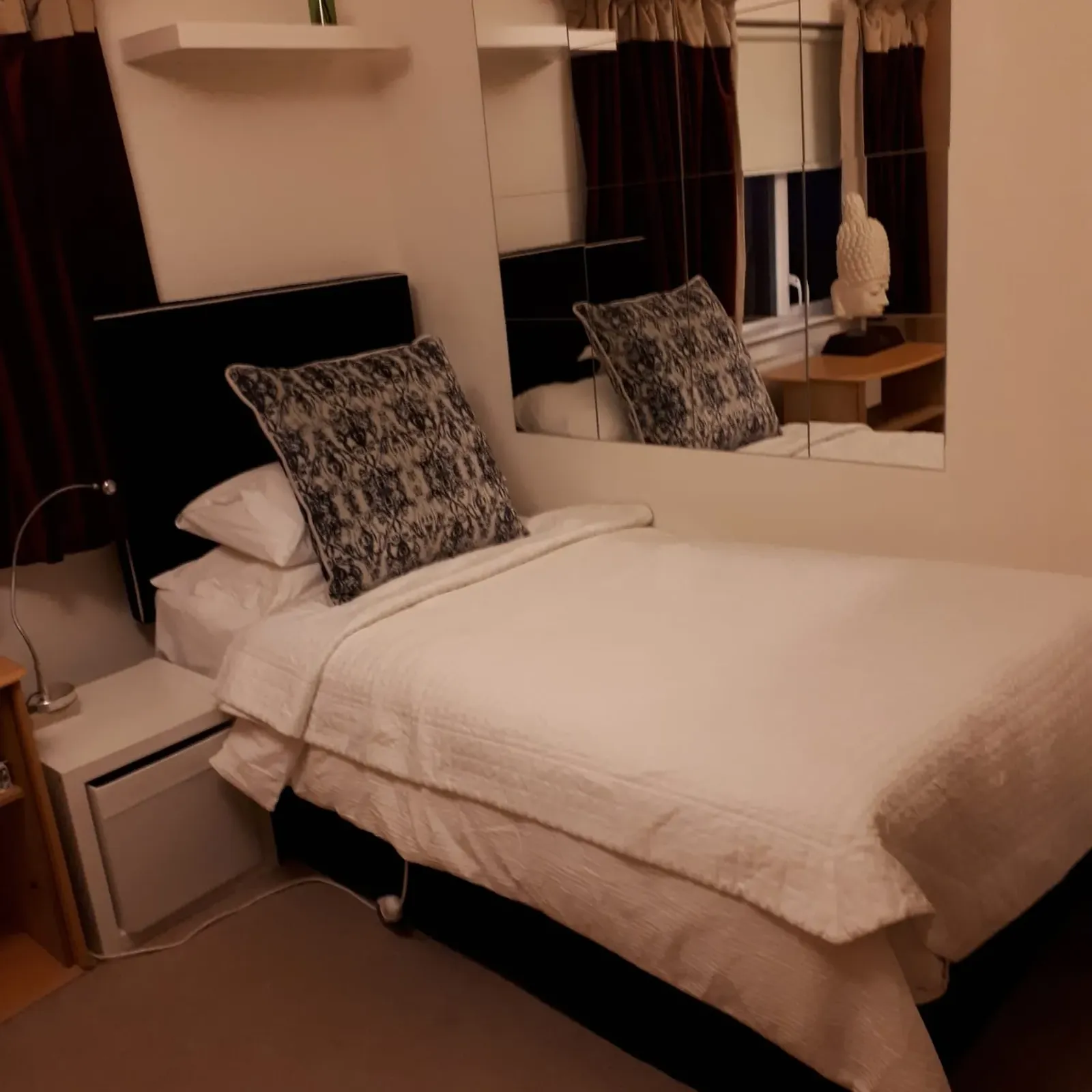 Bedroom with a made bed, two patterned pillows, a mirrored wall above the bedside, curtains and a small bedside table with a decorative bust.