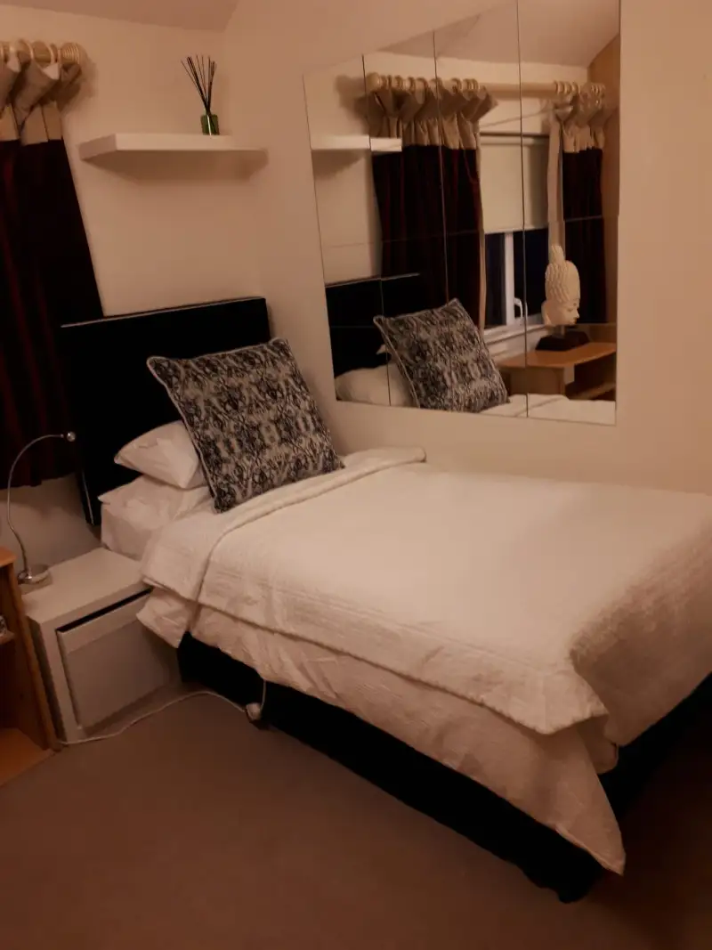 Bedroom with a made bed, two patterned pillows, a mirrored wall above the bedside, curtains and a small bedside table with a decorative bust.