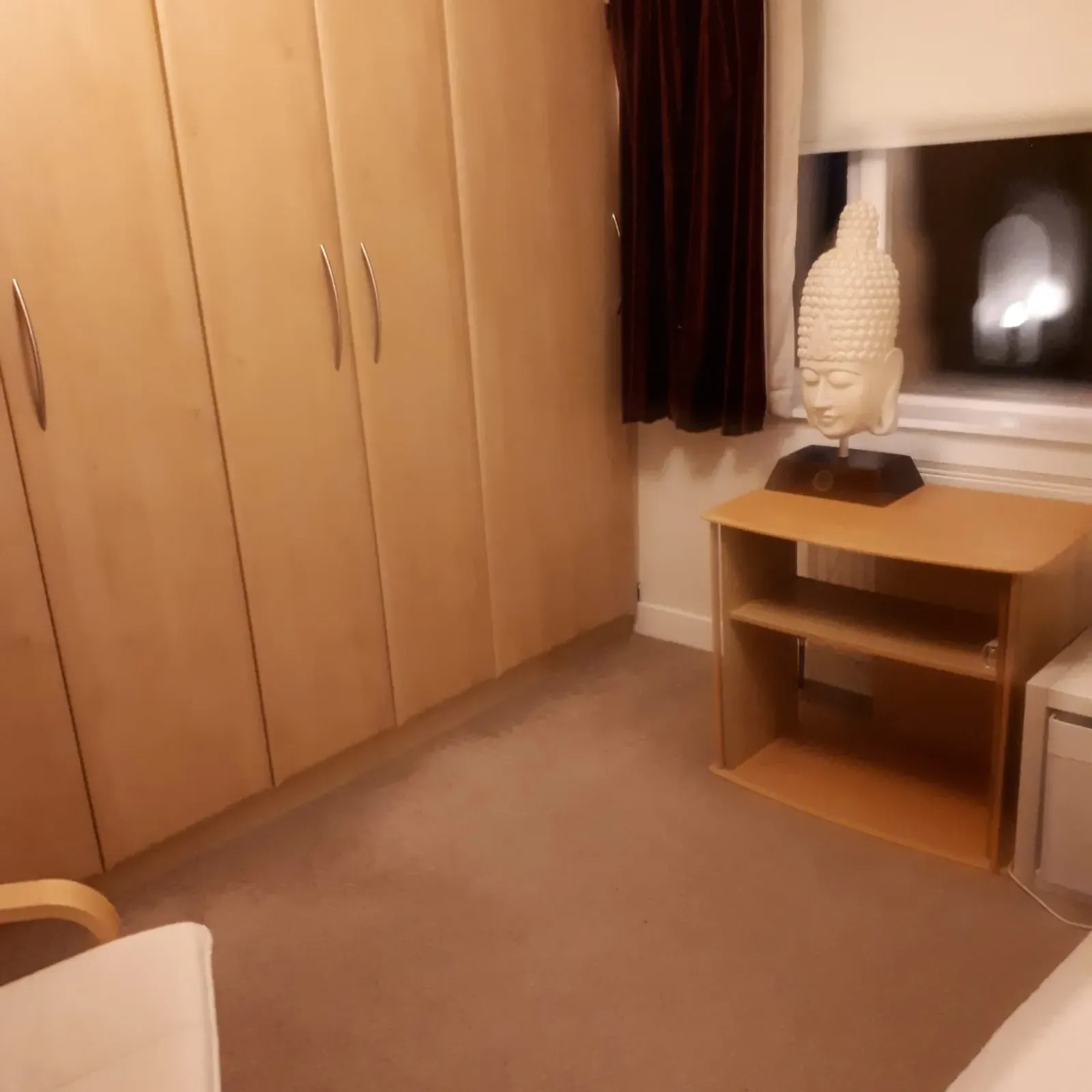 Bedroom corner with built-in wooden wardrobes, a window with dark curtains and a small shelving unit topped by a decorative head statue.