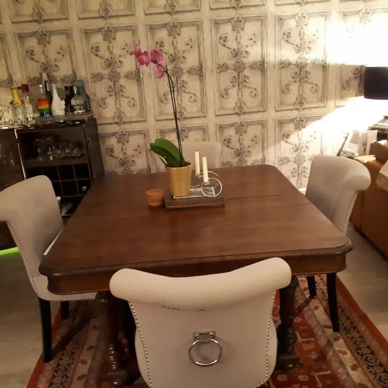 Dining room with a wooden dining table, upholstered chairs, a potted orchid and candlesticks on the table, patterned wall panels, a sideboard with glassware and a floor lamp.