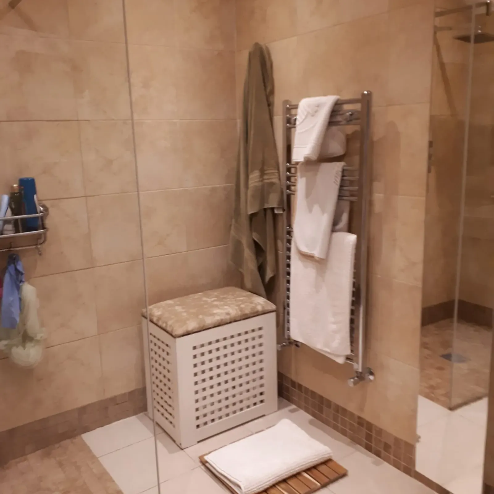 Bathroom with a walk-in shower, glass partition, wall-mounted heated towel rack with folded towels, laundry hamper and bath mat on tiled floor.