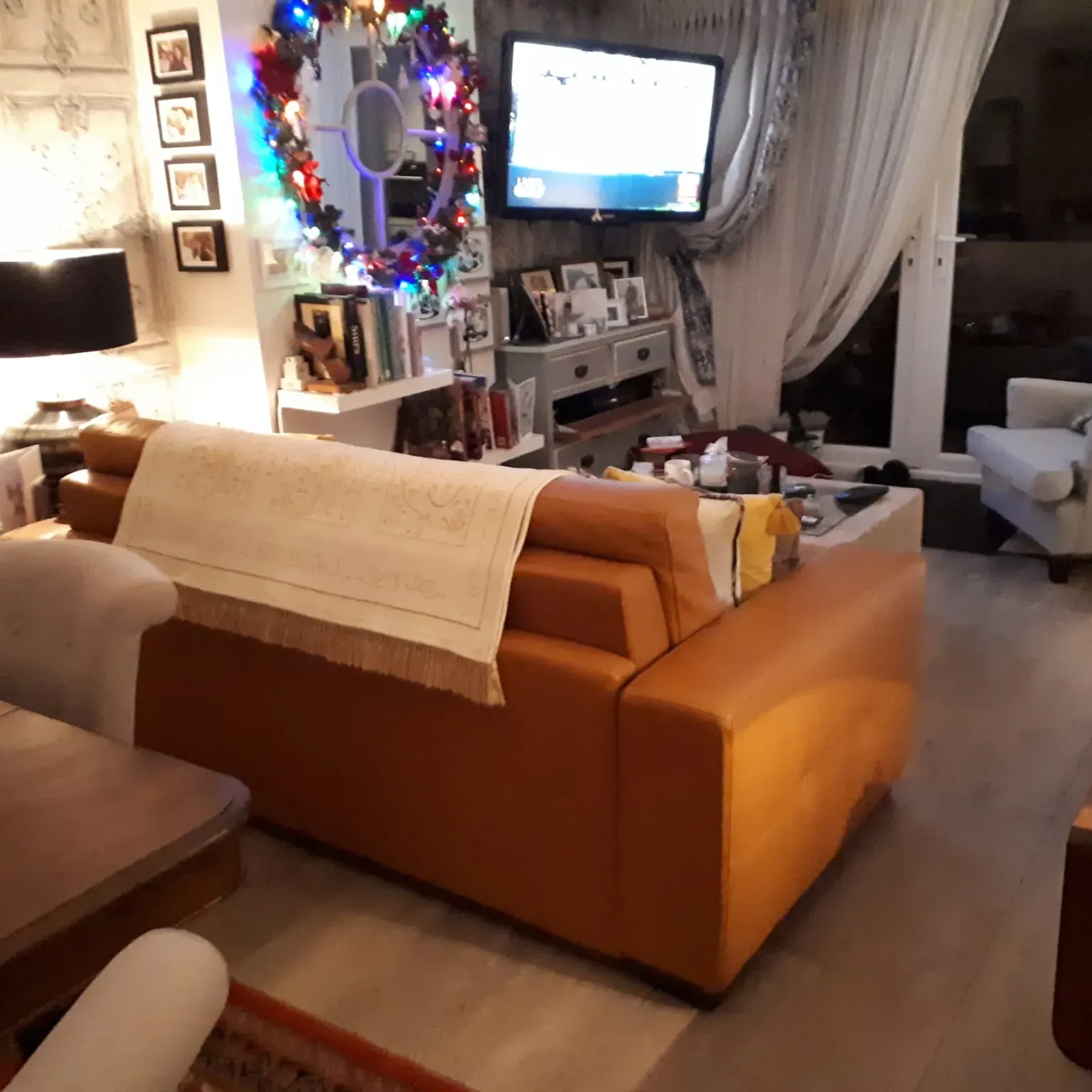 Living room with a tan leather sofa, armchair, wall-mounted TV, coffee table and bookshelf with lit decorative wreath.