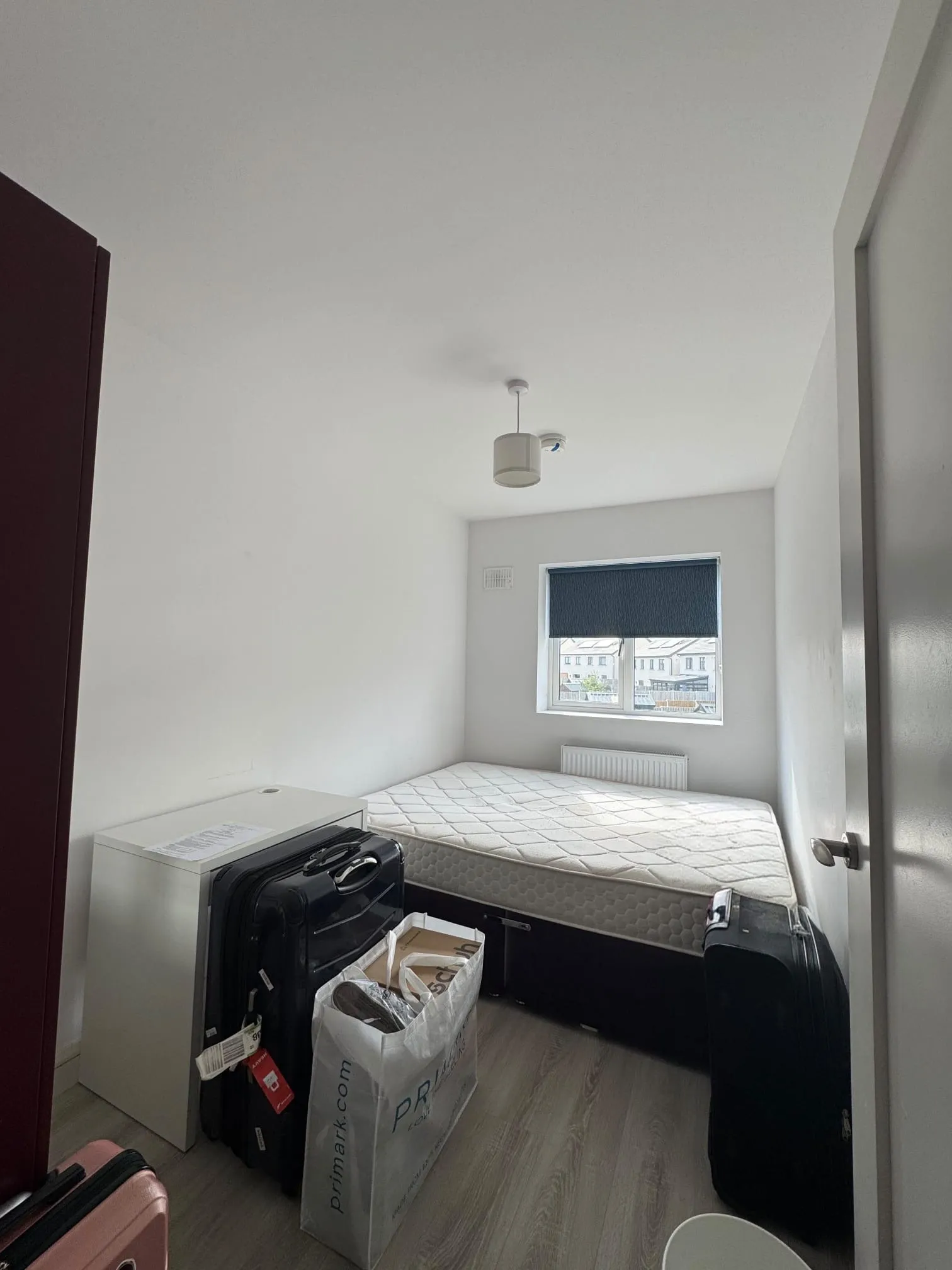 Small bedroom with a bare mattress on a bed frame, a window with a closed blind, radiator below the window and suitcases and bags on the floor.