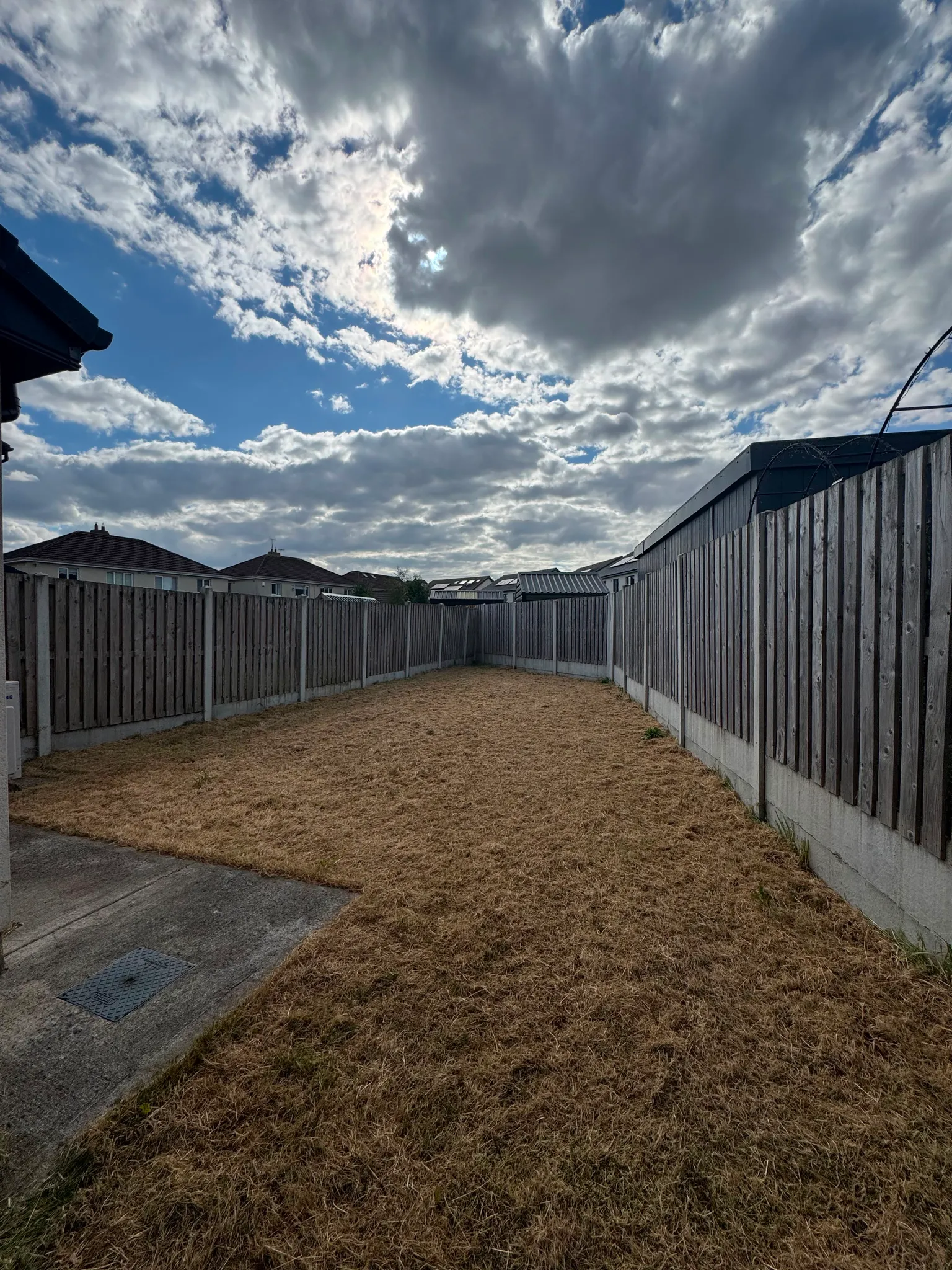 Backyard with a fenced perimeter, dry grass lawn, concrete path at left and cloudy sky above.