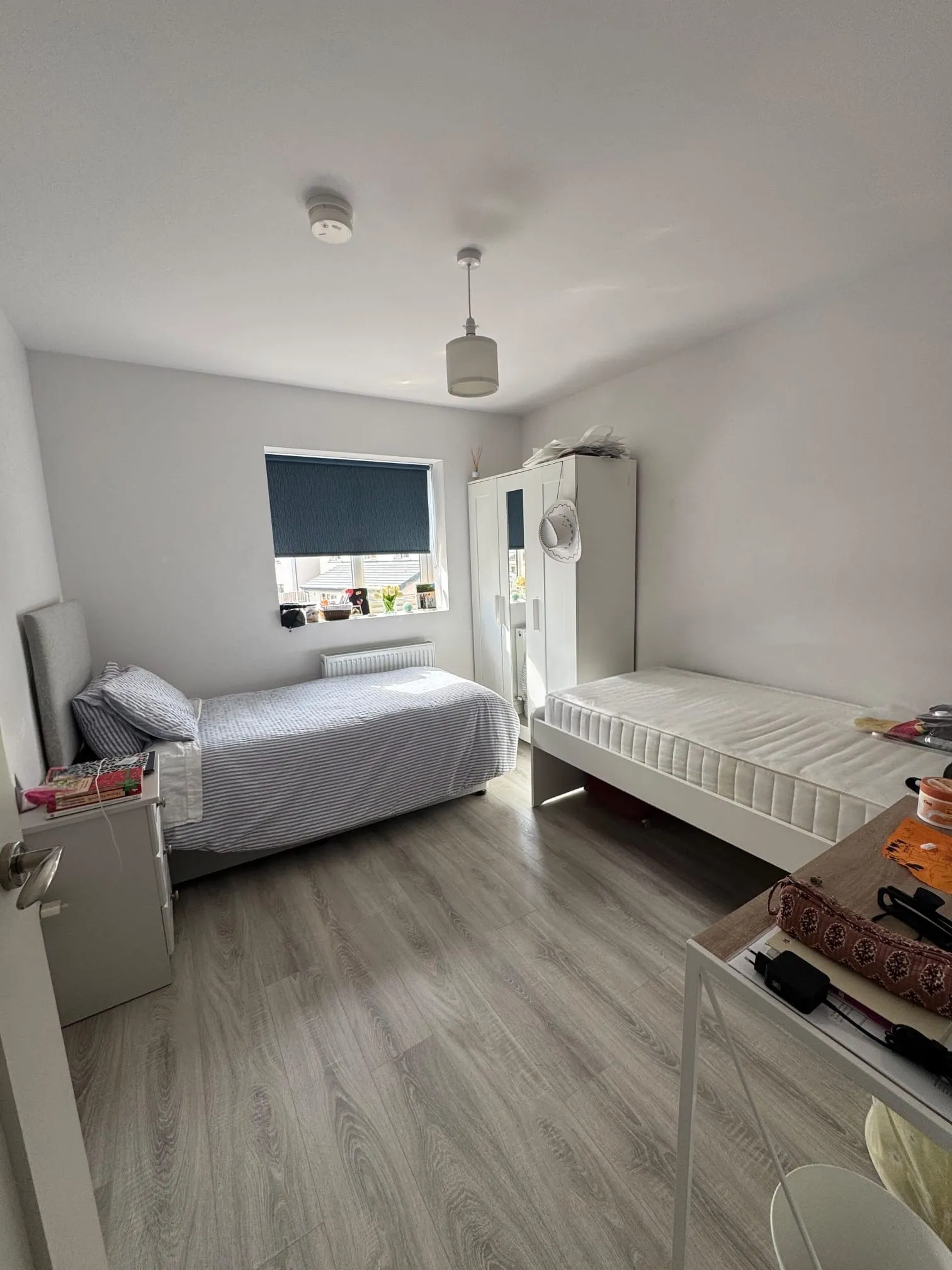 Bedroom with two single beds, a wardrobe, a window with a blind, bedside table and overhead pendant light.