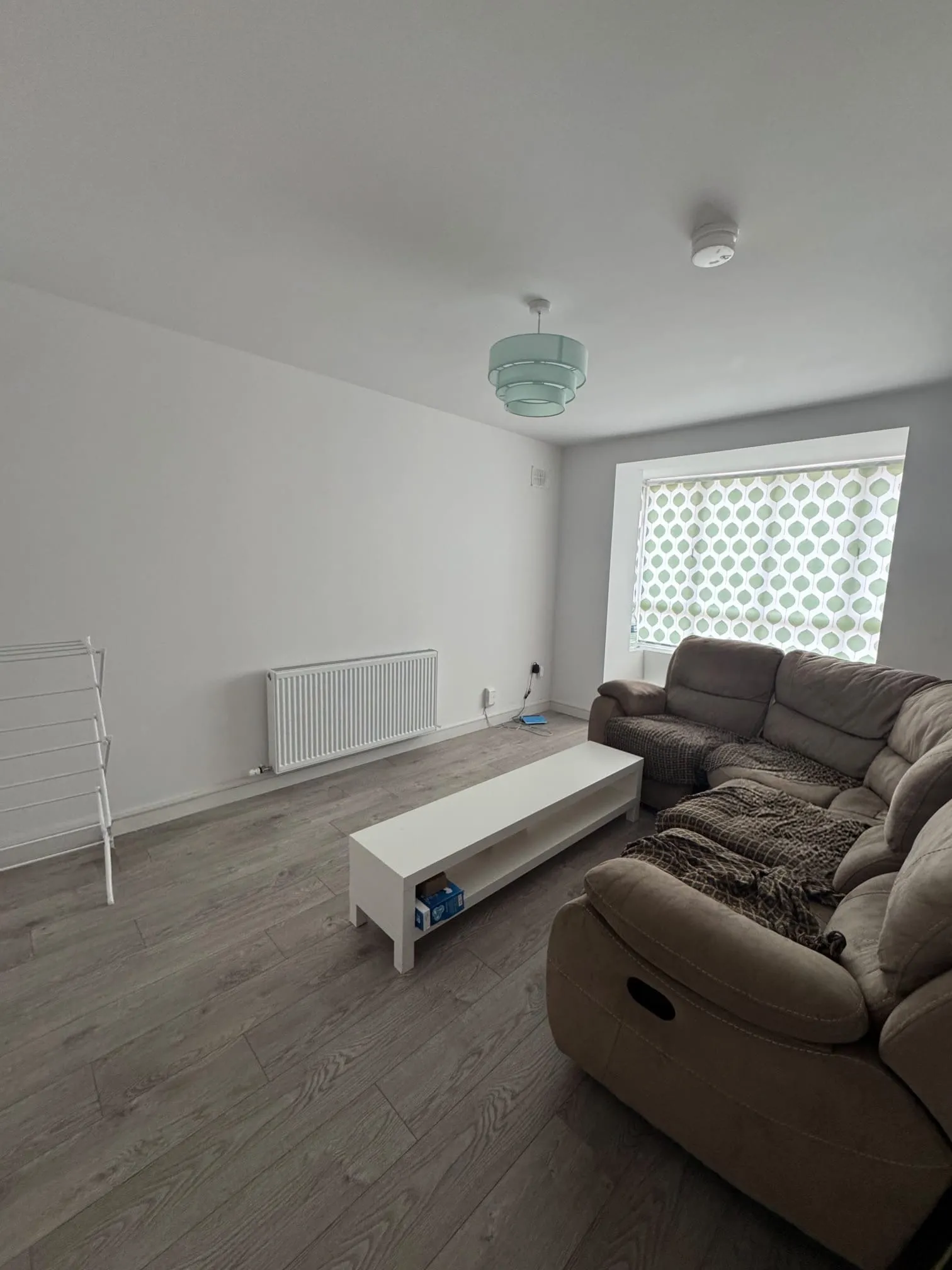 Living room with a beige sofa, low white coffee table, radiator on the wall and a window with a patterned roller blind.