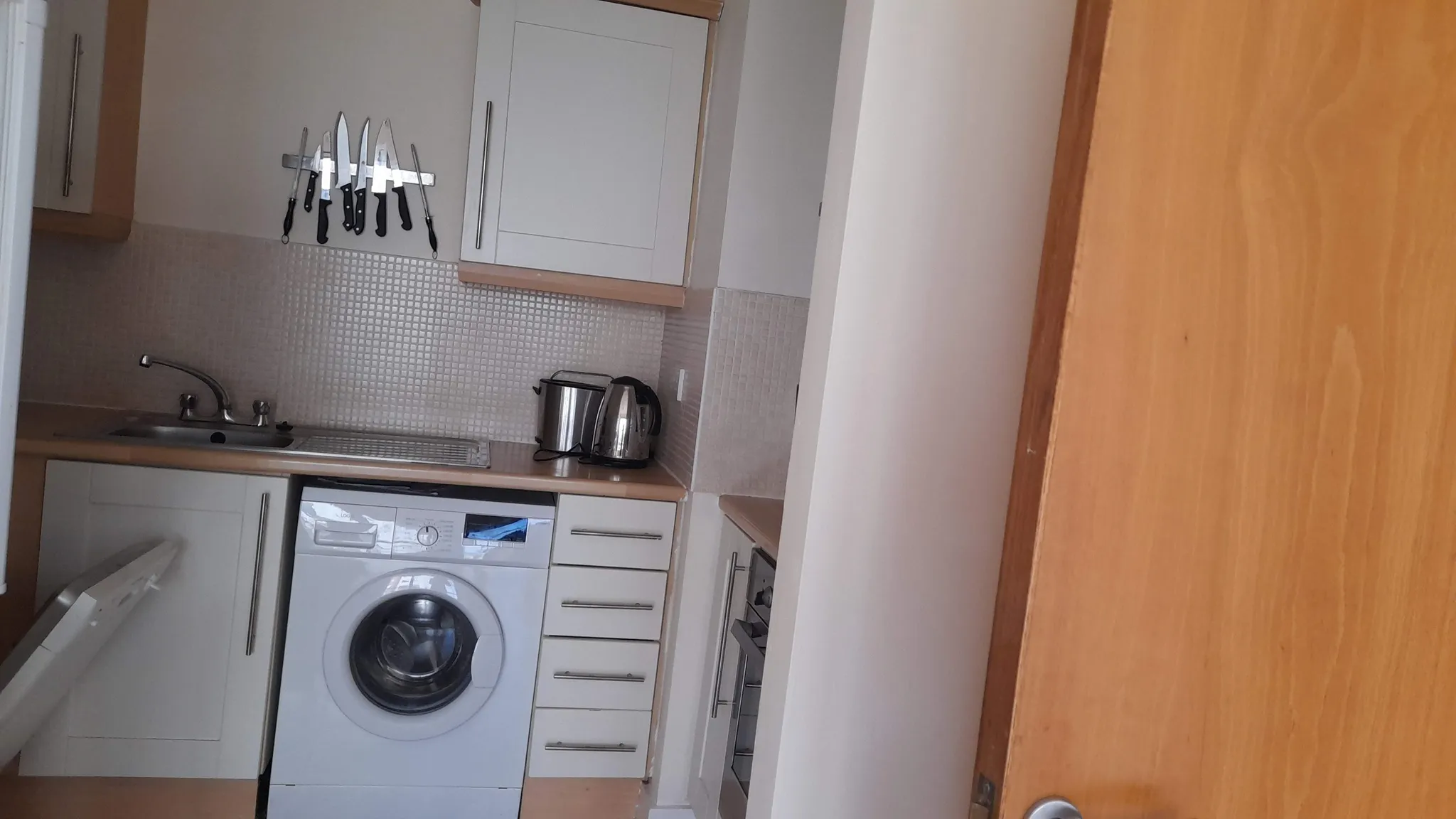 Small kitchen with sink, front-loading washing machine, countertop kettle and bin, white cabinets and a magnetic knife strip on the wall.
