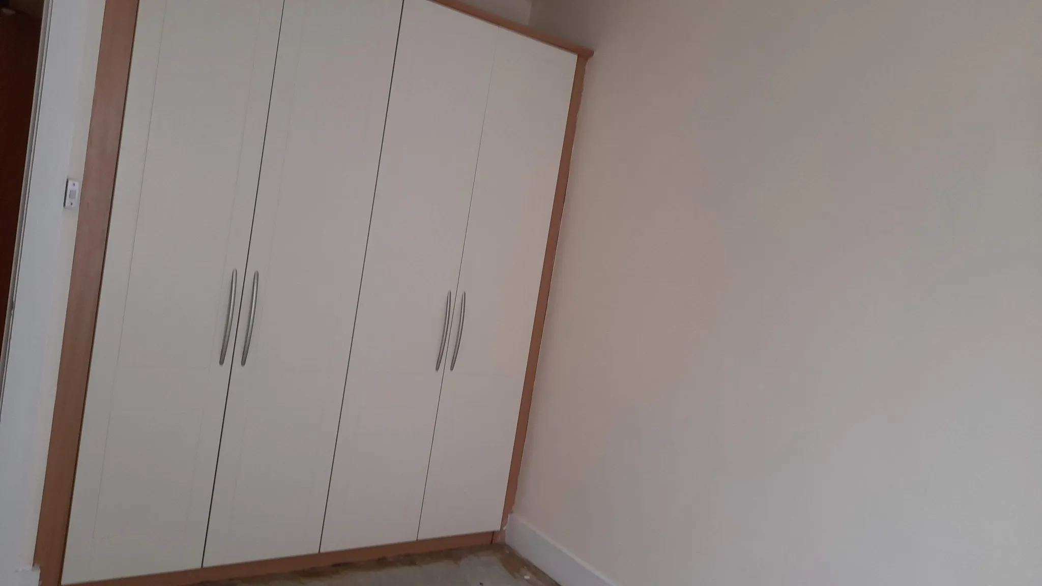 Bedroom corner showing a built-in white wardrobe with vertical handles and an adjacent blank wall.