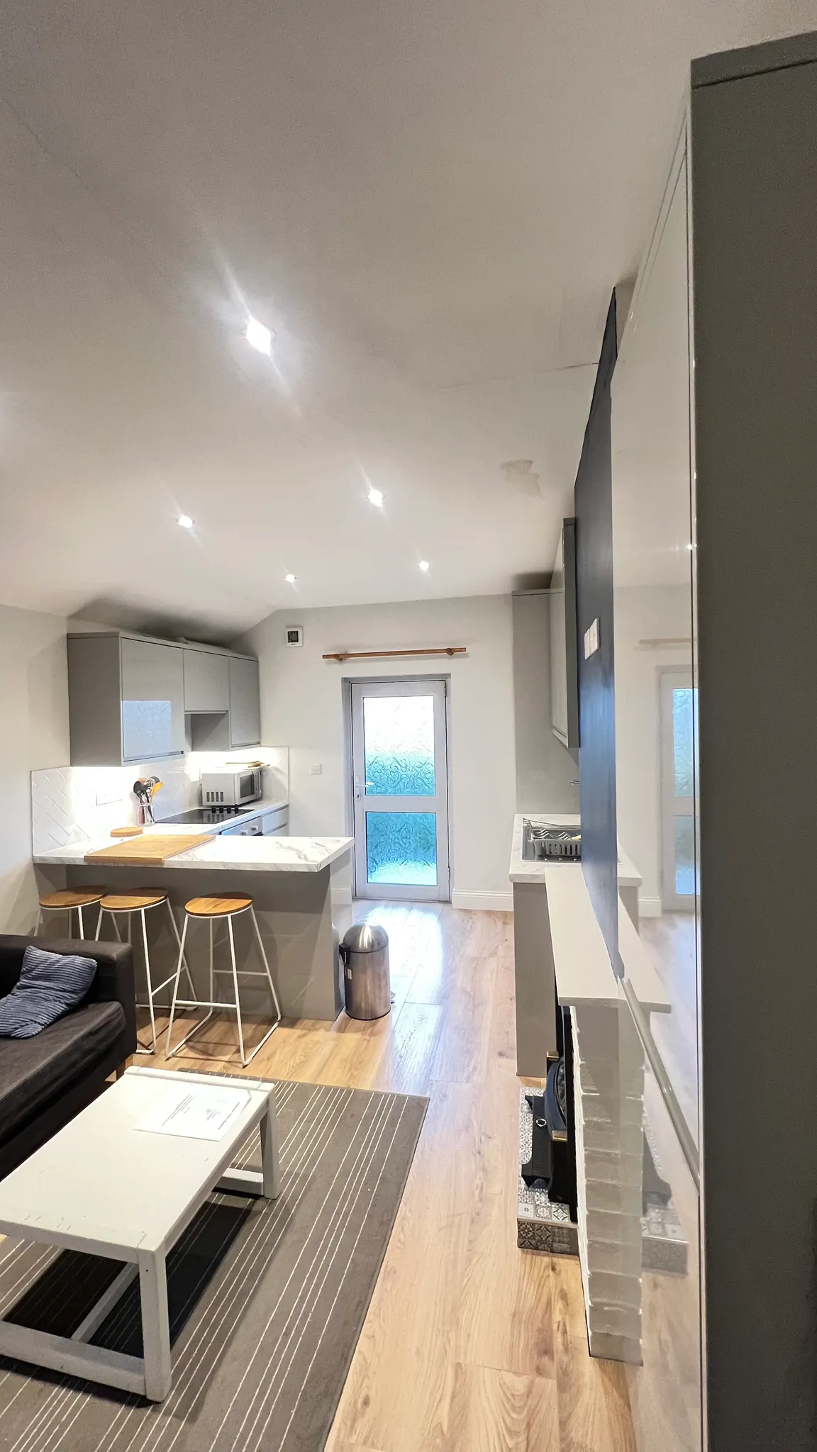 Open-plan living area with a small kitchen island and three stools, sofa and coffee table, recessed ceiling lights, and a frosted glass door at the back.