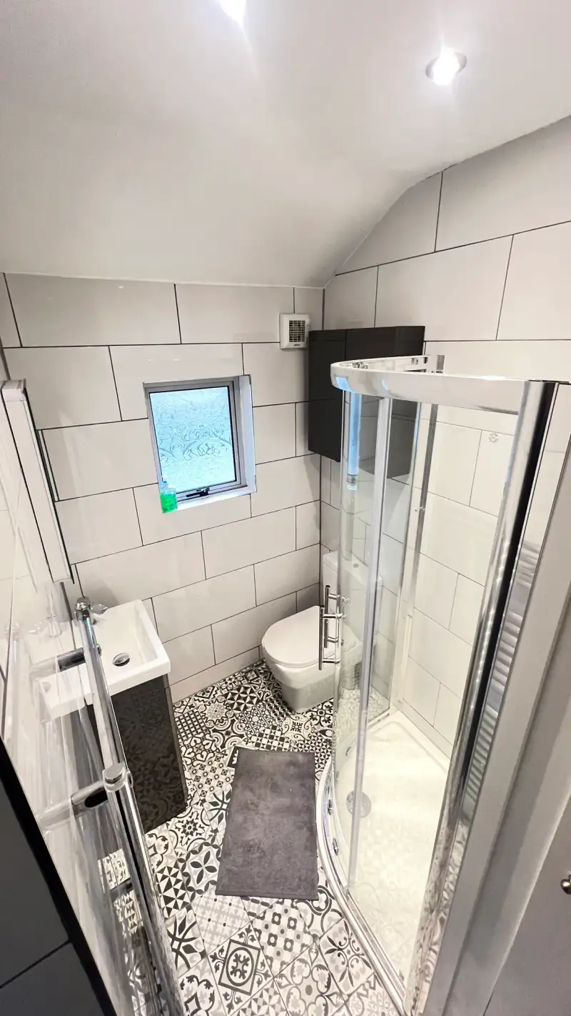 Bathroom with a corner glass shower, toilet, small sink with vanity, patterned floor tiles and a small frosted window.