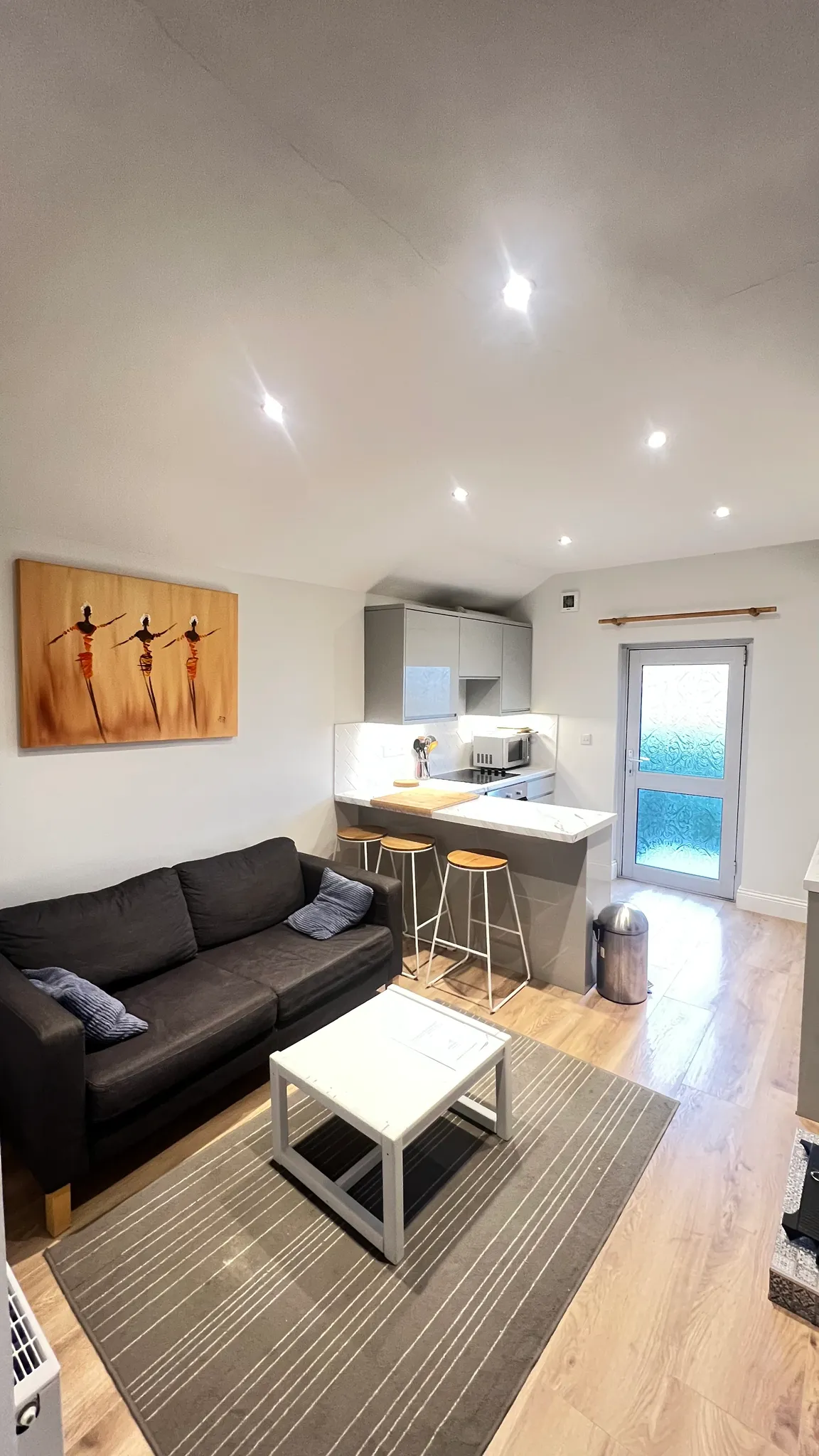 Open-plan living room and kitchen with a dark sofa, coffee table on a striped rug, kitchen island with three bar stools, wall artwork and a back door with frosted glass.