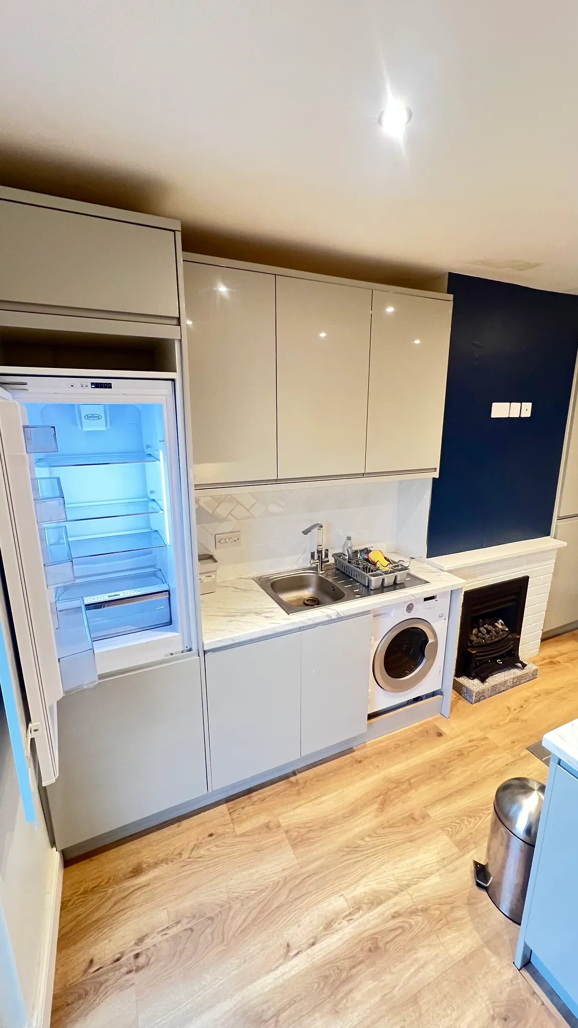 Kitchen with upper cabinets, open refrigerator, sink with dish rack, front‑loading washer, small fireplace and wood floor.