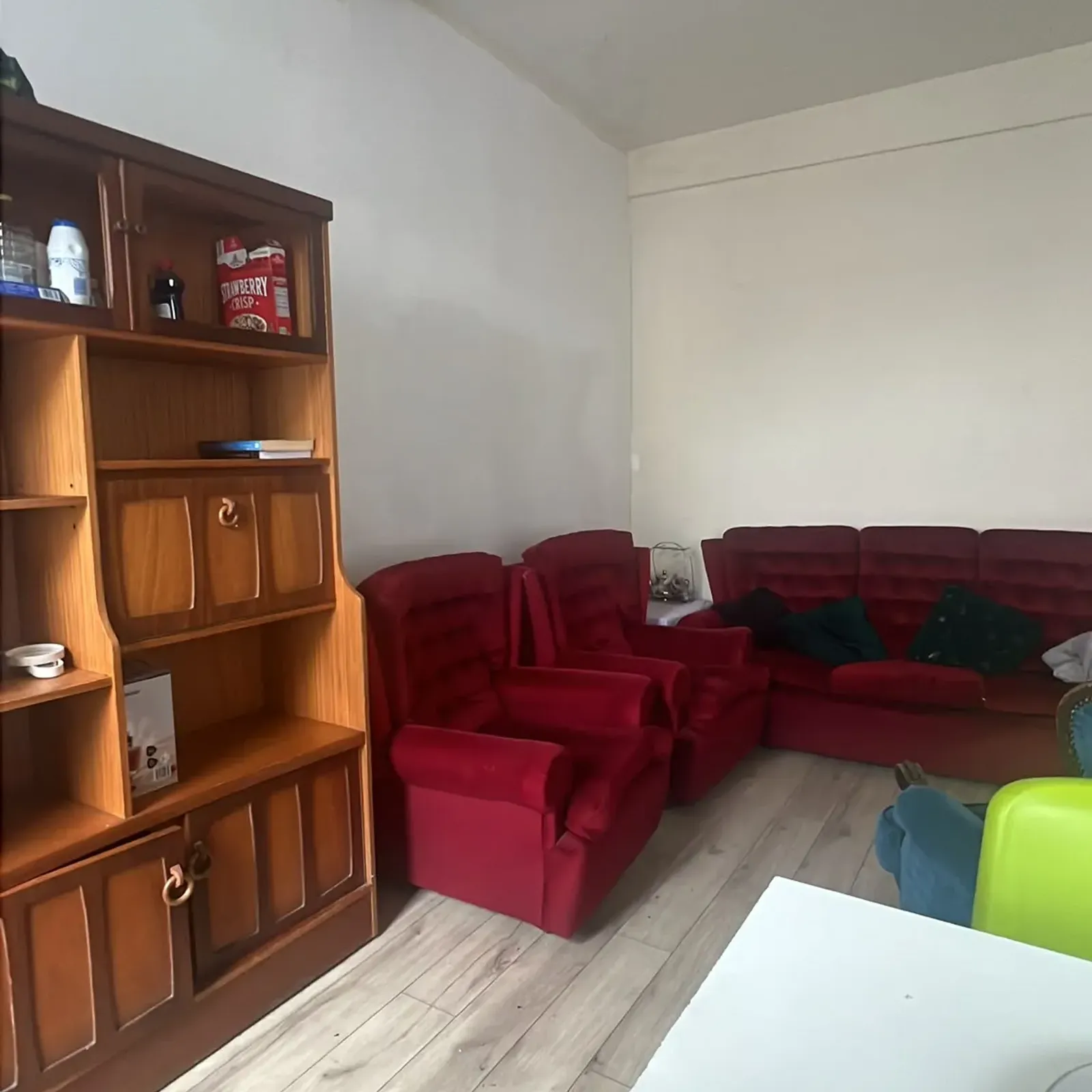 Living room with red upholstered armchairs and a matching sofa, a wooden shelving unit, light wood laminate floor and a visible ceiling beam.
