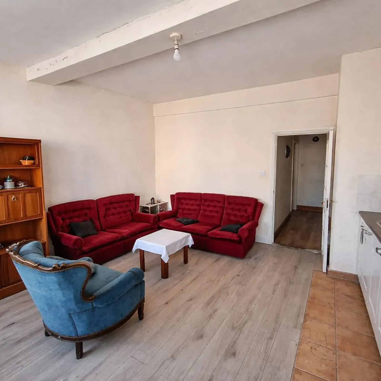 Living room with two red sofas, a blue armchair, small central table, wooden bookshelf, and a kitchenette with sink and washing machine along the right wall.
