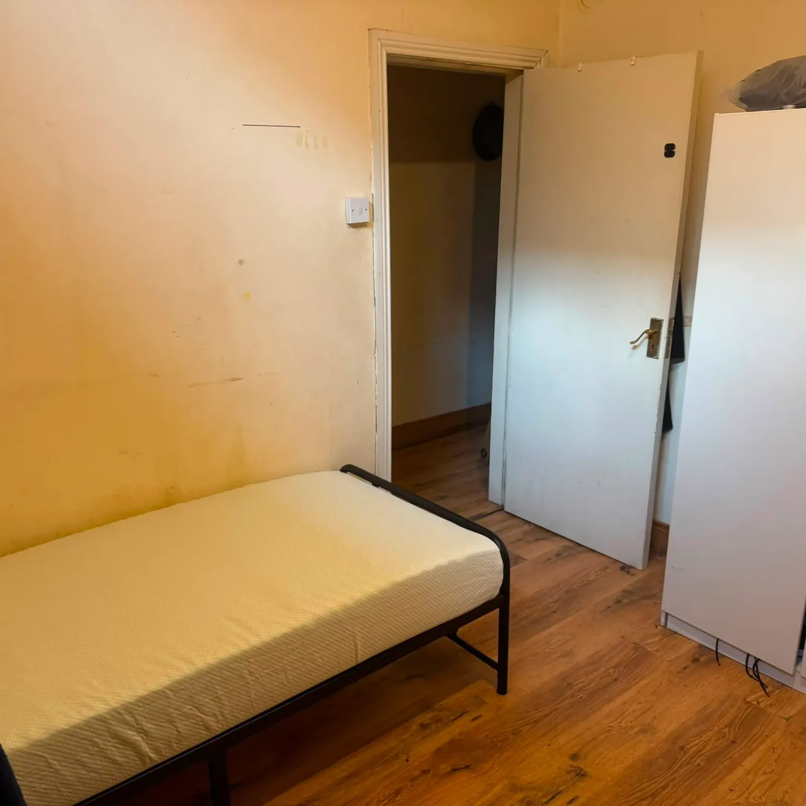 Bedroom with a single bed on a metal frame, an open door leading to a hallway, and a white wardrobe on a wooden floor.